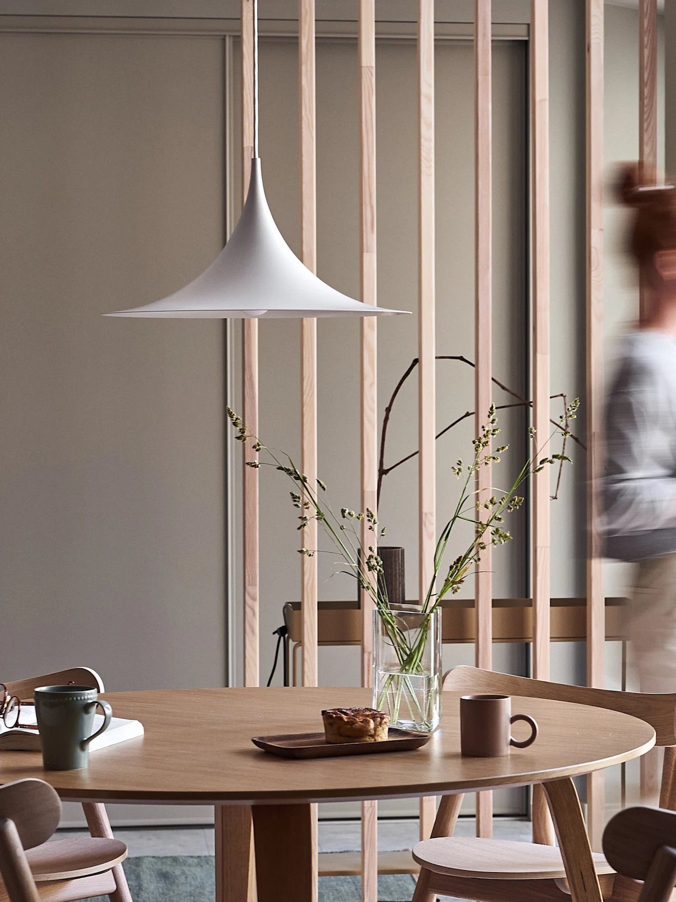 Modernt matplats med runt träbord, vit pendellampa, vas med gräs, bakverk och muggar, med en suddig person som går förbi.