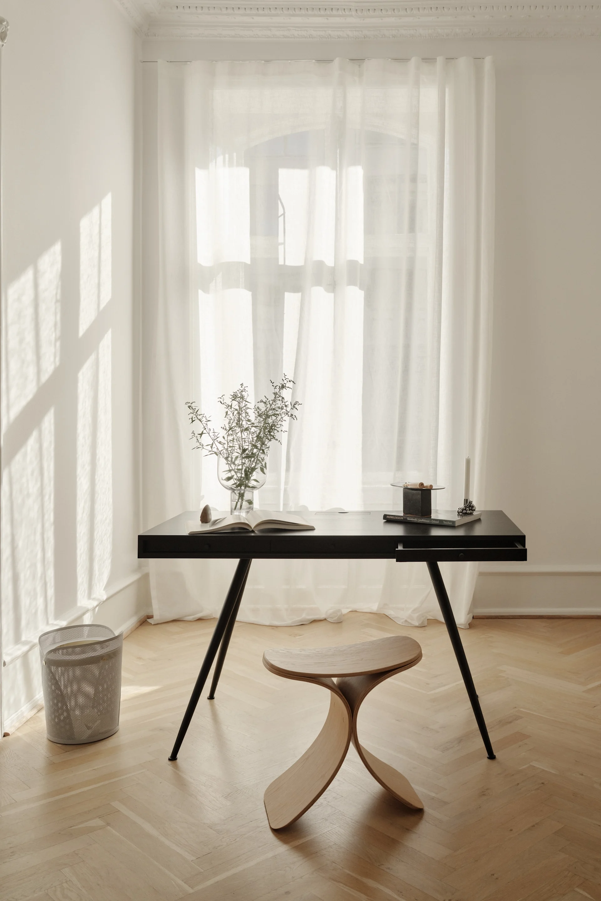 Ett minimalistiskt hemmakontor med ett svart skrivbord, en böjd träpall, en vas med grönska och en öppen bok vid ett fönster med skira gardiner.