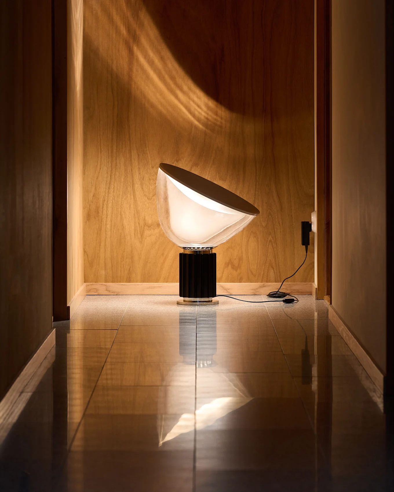 Modern lampa med halvcirkelformad, vit skärm och svart räfflad bas i träpanelad hall med reflekterande golv. Ljusmönster på väggen.