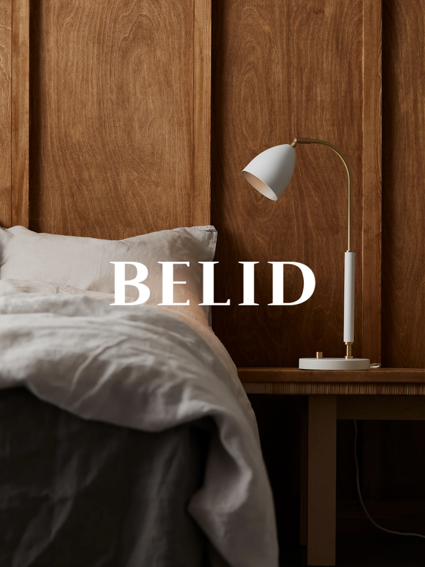 Vit och mässingsfärgad lampa på ett nattduksbord i trä, med texten 'BELID' över en säng med lakan i linne.