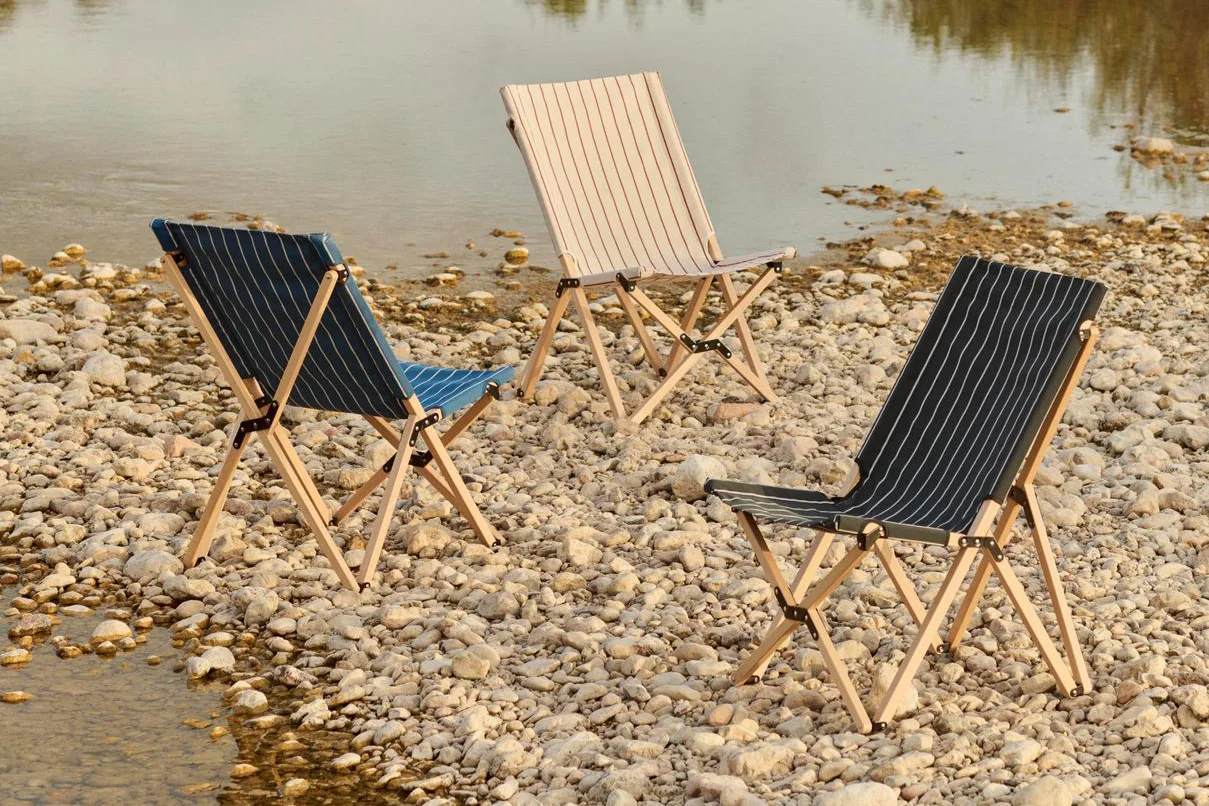 Tre randiga fällbara campingstolar på en stenig strand vid vattnet: blå, beige och mörkgrå.