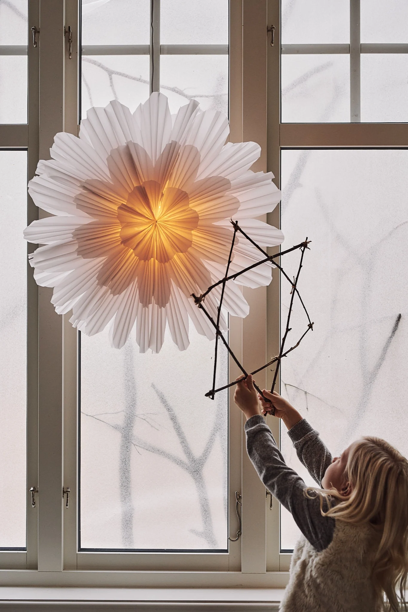 Den sagolika adventsstjärnan Snöblomma från Watt & Veke hänger i fönstret medan barnet står bredvid med sin egengjorda julstjärna av pinnar.