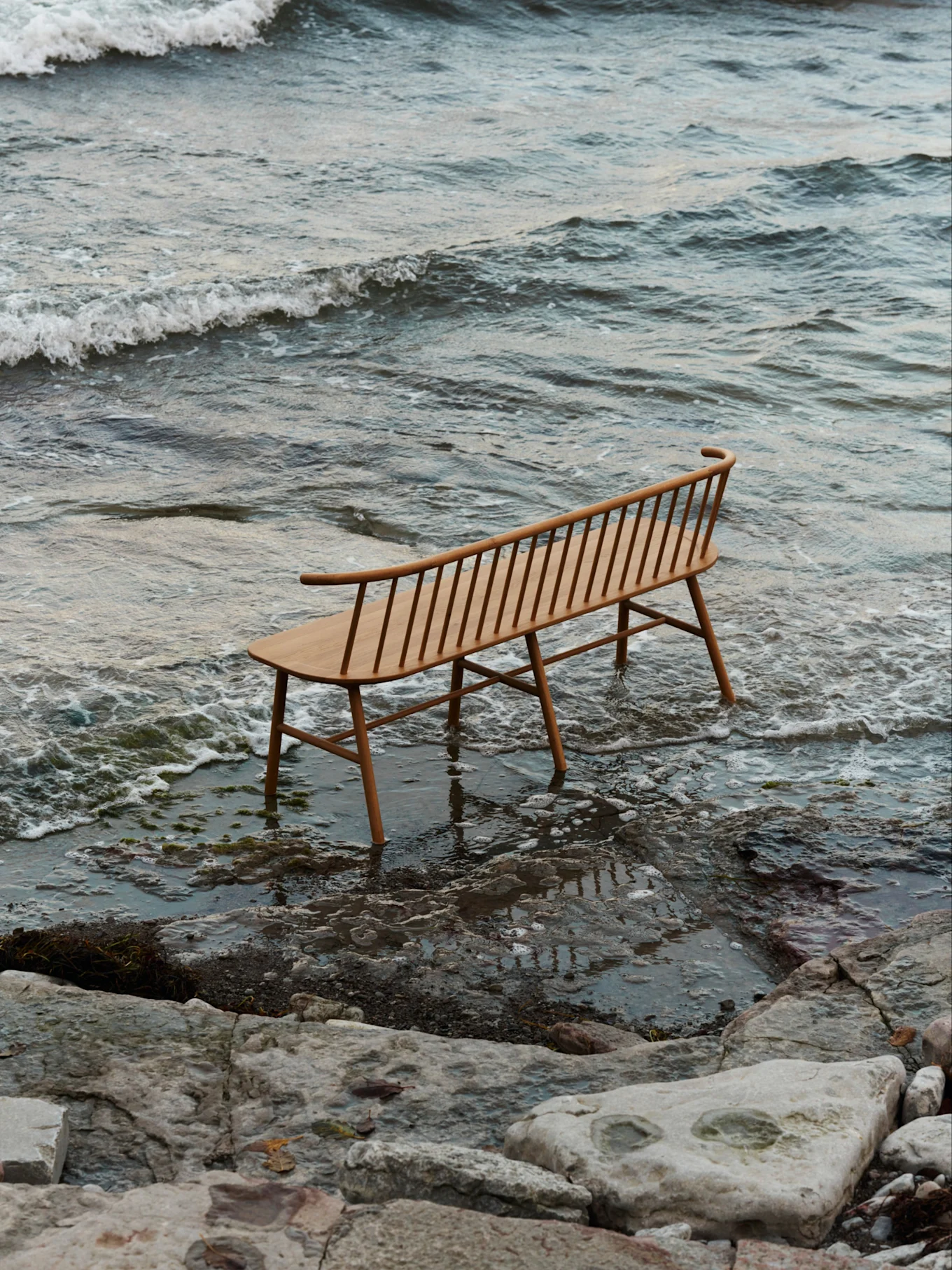 En ljus träbänk med ribbor och böjt ryggstöd står i grunt vatten på en stenig strand med milda vågor.