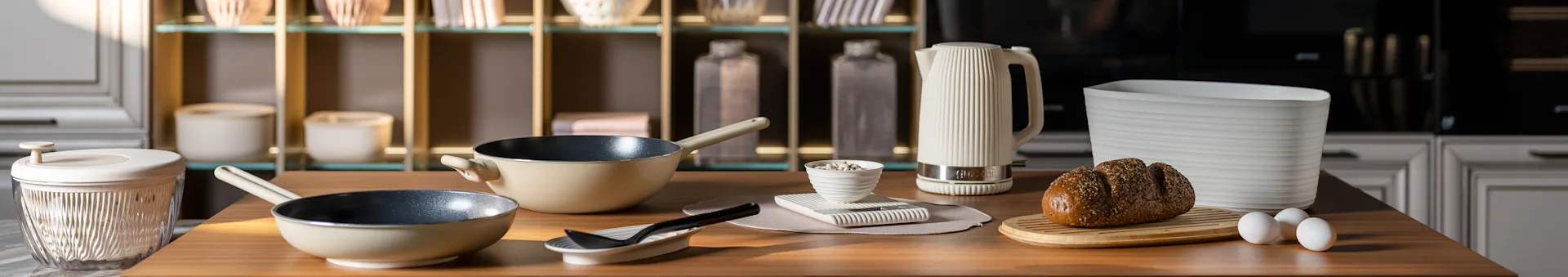 Köksbänk med beiga stekpannor, en vit räfflad vattenkokare, ett fröbröd på en skärbräda och ägg. Moderna köksaccessoarer.