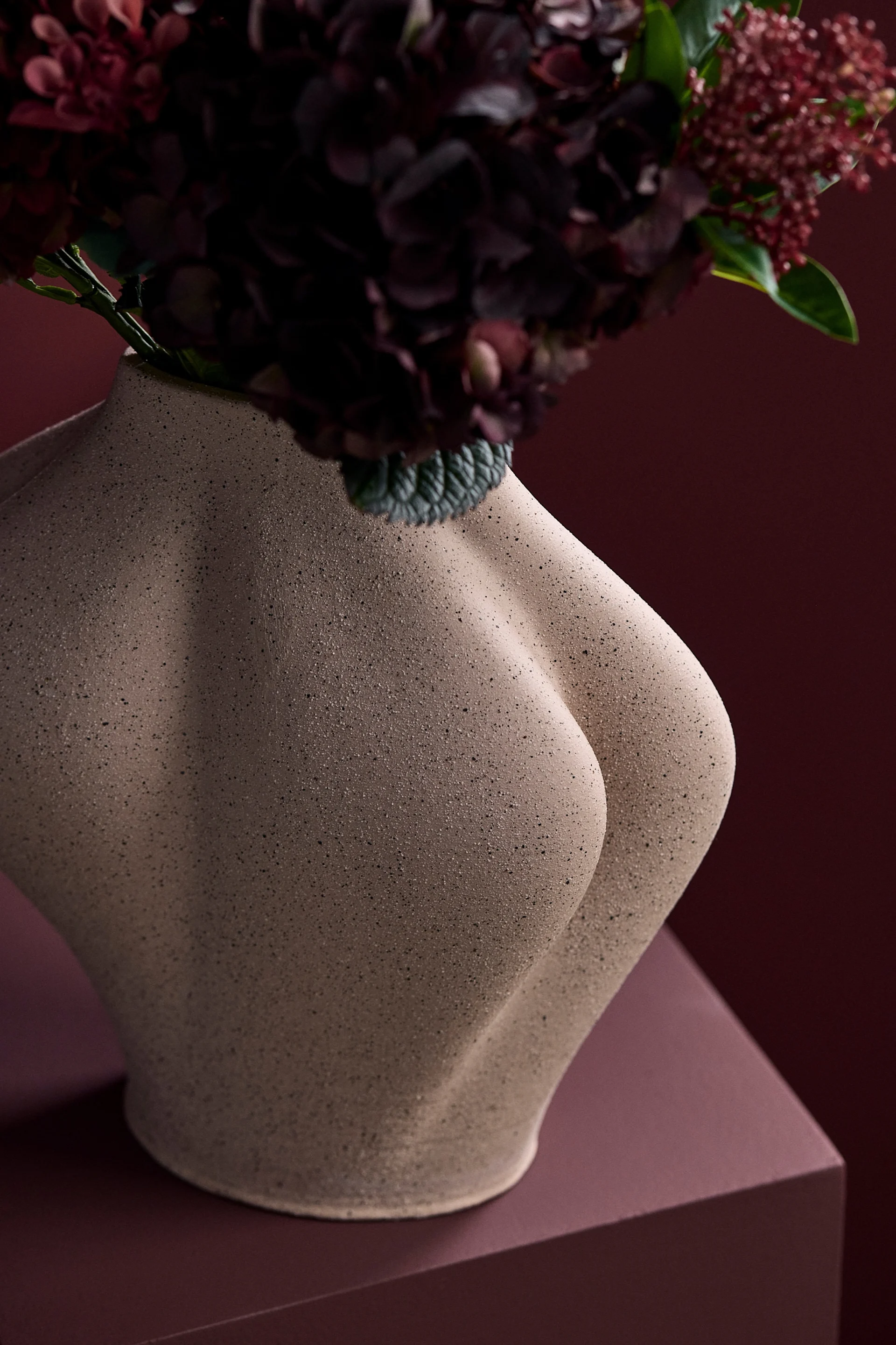 Skulptural beige vas formad som en mänsklig torso, med mörkröda blommor mot en matchande röd bakgrund.