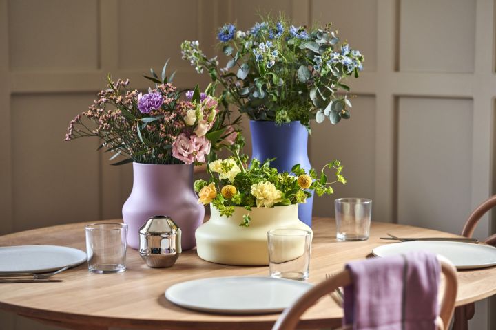 De nya Dorotea-vaserna från Gense, formgivna av Monica Förster. Här ser du en gul låg vas, en syrénlila vas och en hög kleinblå vas med matchande buketter i vasernas färger. Placerade på ett bord med en enkel dukning.