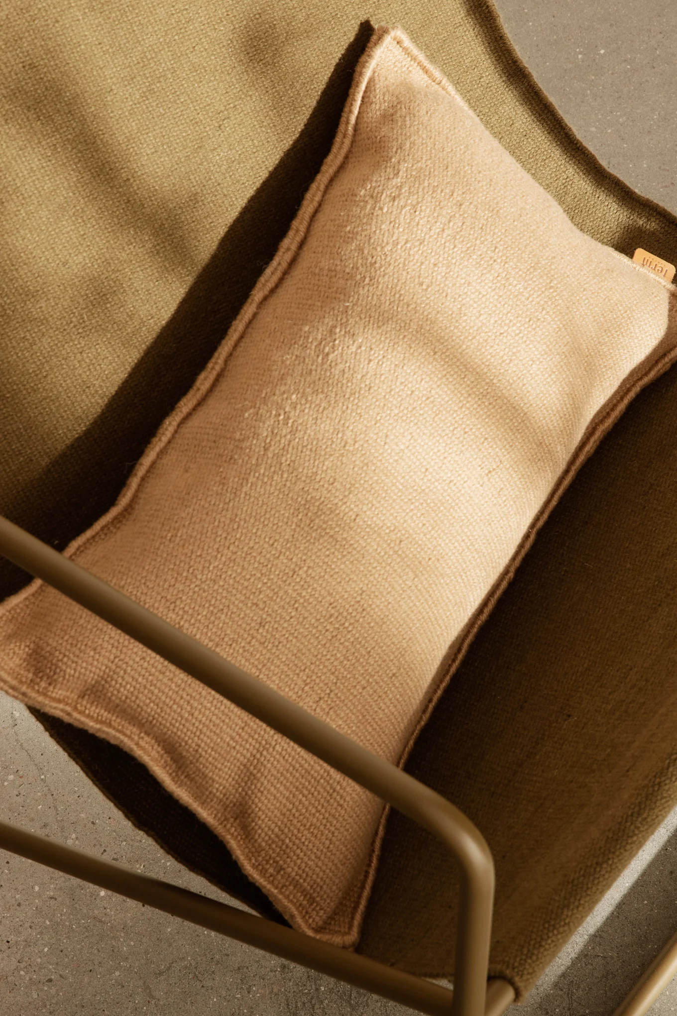 Ovanifrån-bild av en ljusbrun kudde på en mörk olivgrön texturerad tygstol, med del av en metallram och betonggolv.