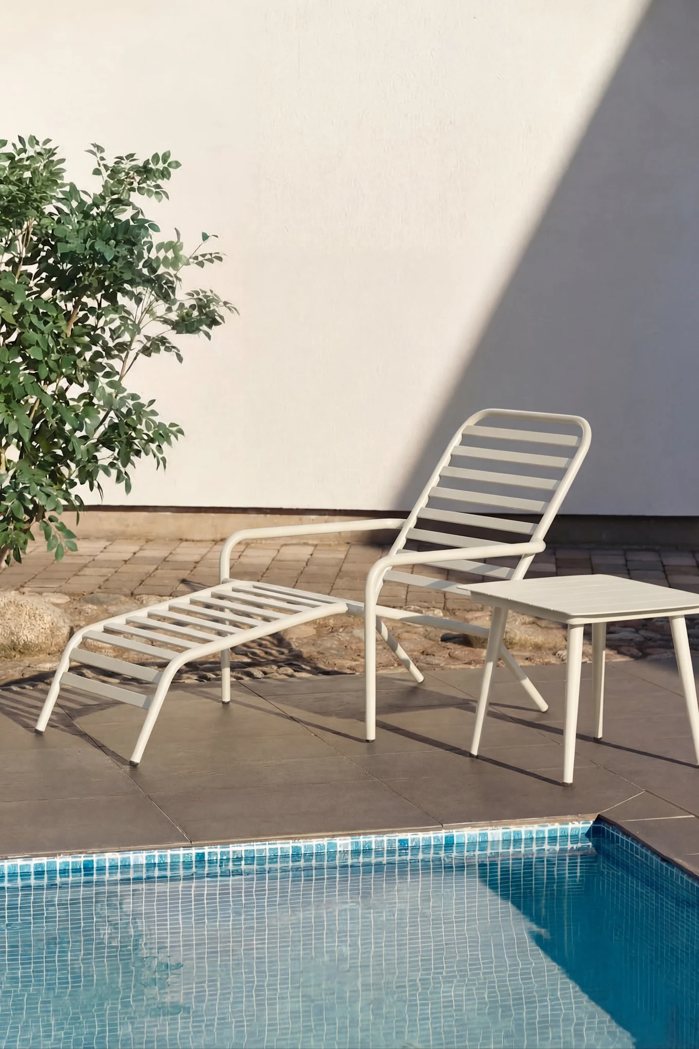 Vit solstol och litet vitt sidobord vid en pool med turkost vatten. Gröna växter och en vit vägg i bakgrunden.