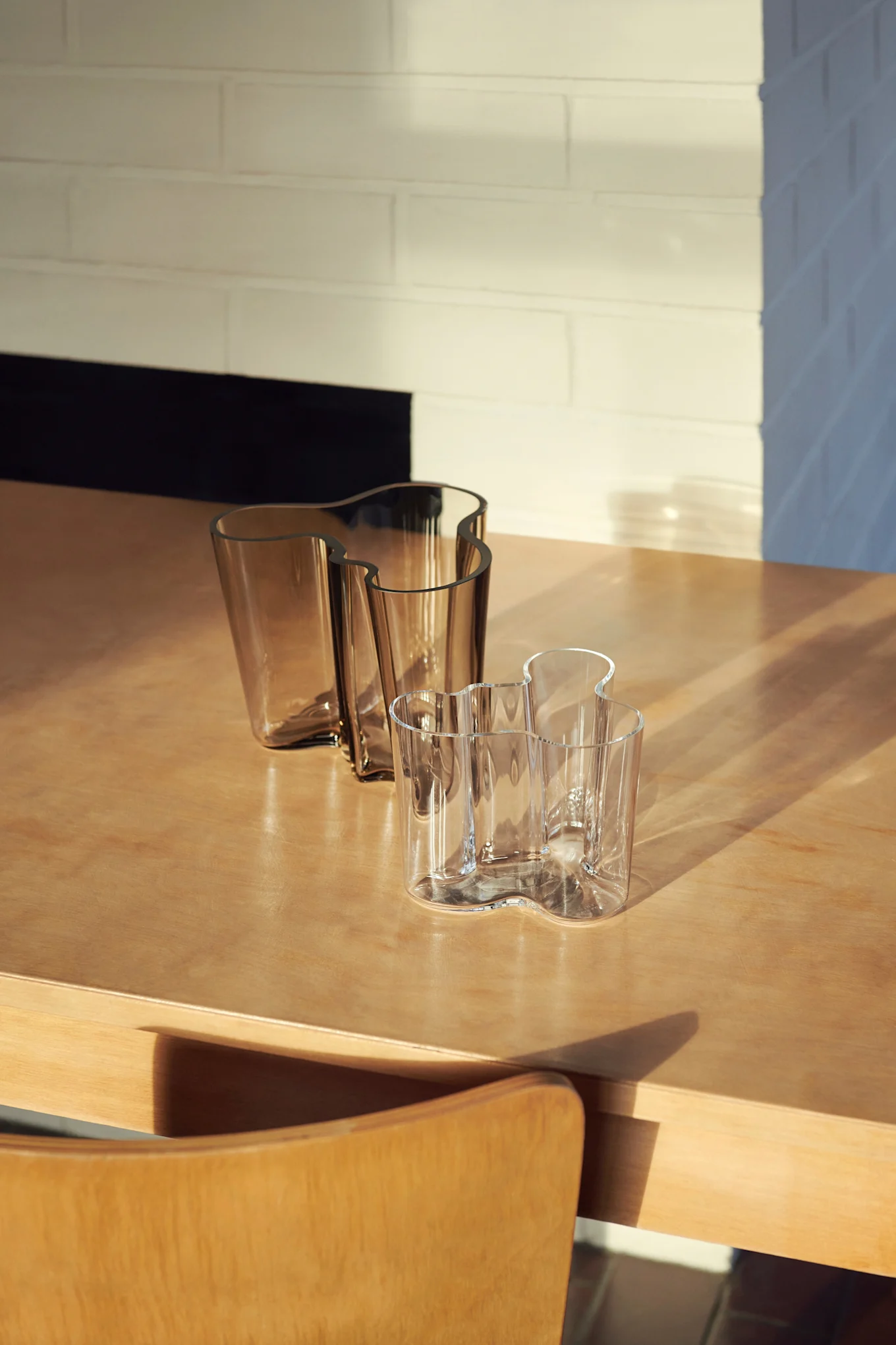 Tre vågiga glasvaser på ett träbord. Två är bärnstensfärgade, en är klar, med solljus och skuggor över scenen.