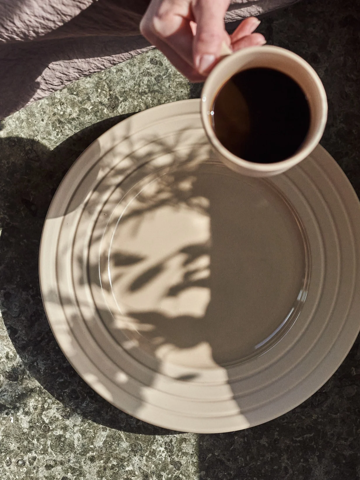 Hand håller kaffekopp över beige tallrik med bladskuggor på ett fläckigt bord.