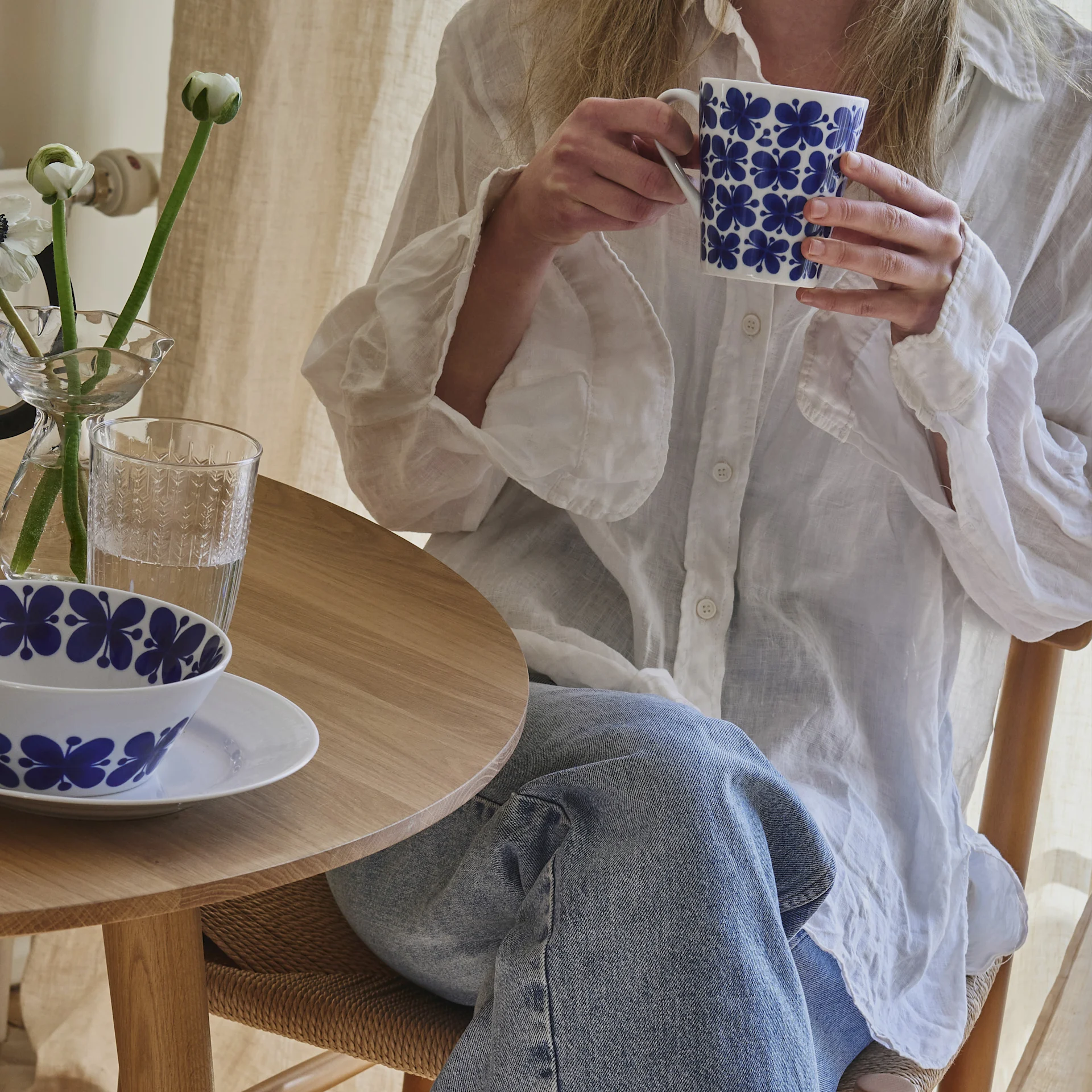 Person i vit linneskjorta och jeans håller en vit mugg med blått blommönster, sitter vid ett träbord med en matchande skål och blommor.