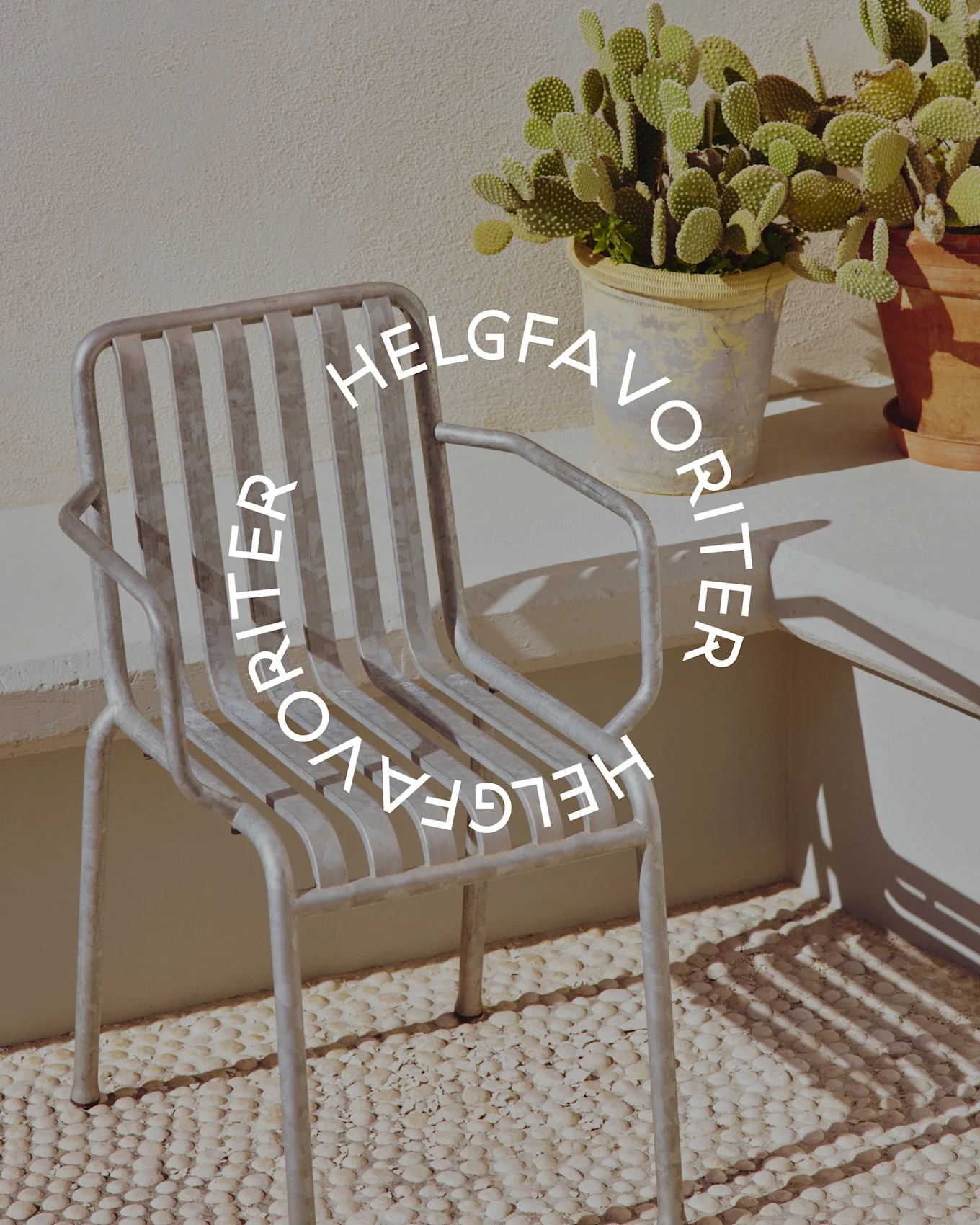 Ljusgrå metallstol med texten 'Helgfavoriter', på en småstensbelagd yta intill en kaktus i kruka.