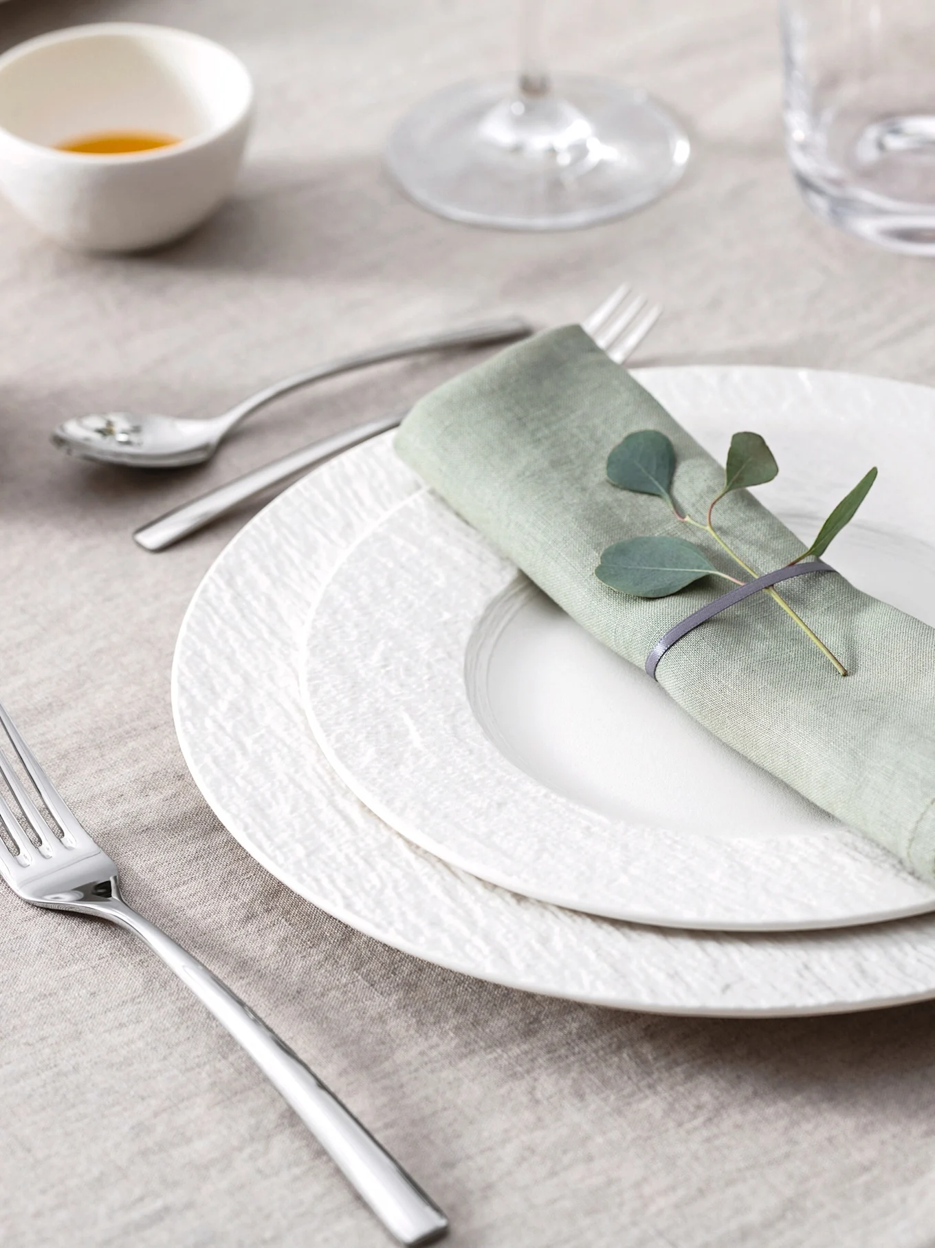 Elegant dukning med ljusgrön linneservett, eukalyptusblad, vita texturerade tallrikar och bestick på en beige bordsduk.