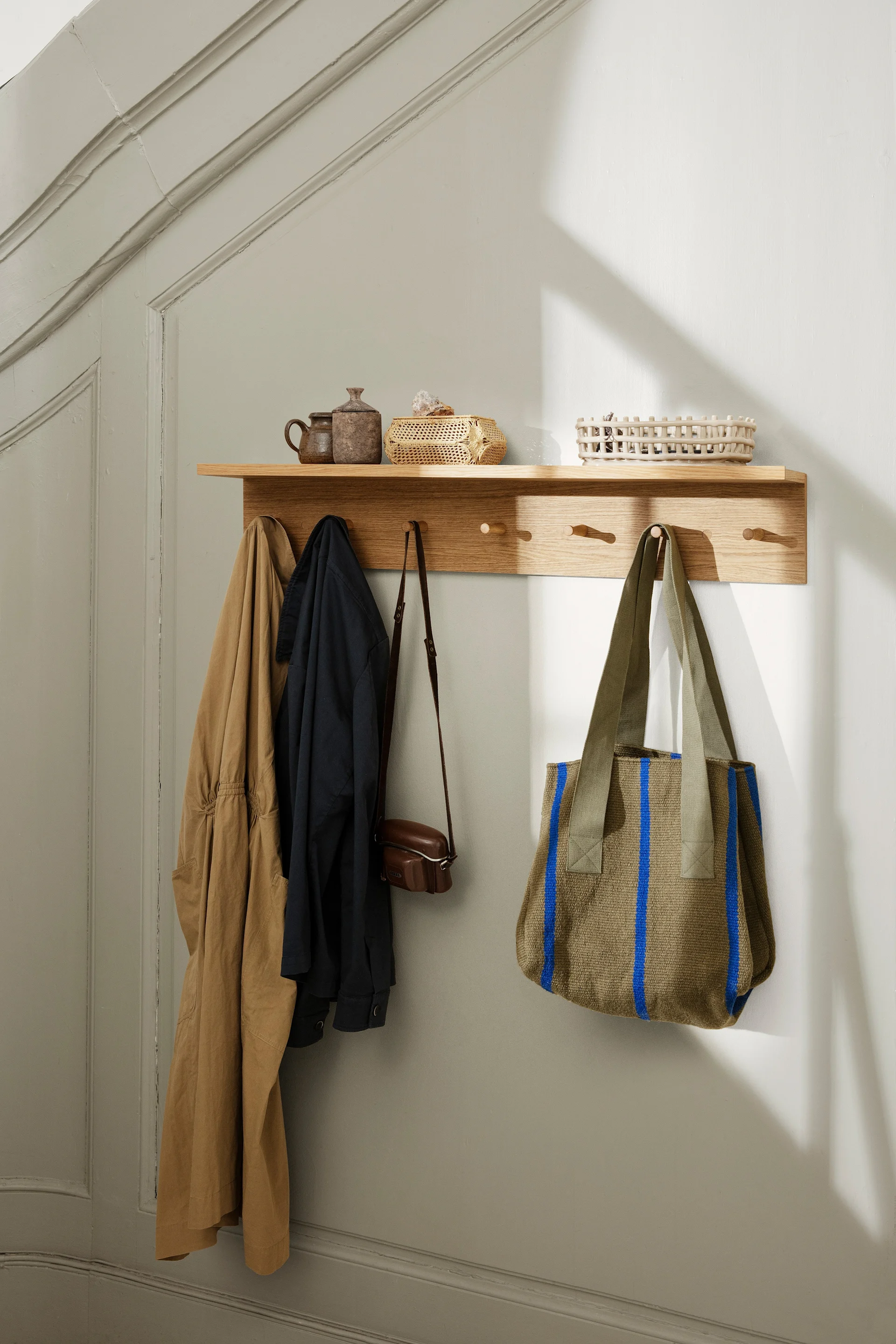 Place Rack knopplist hylla i trä med krokar från Ferm Living. Två jackor, liten läderväska och olivgrön tygväska med blå ränder. På hyllan: krukor och korgar. En hylla är perfekt för att dekorera vägg.