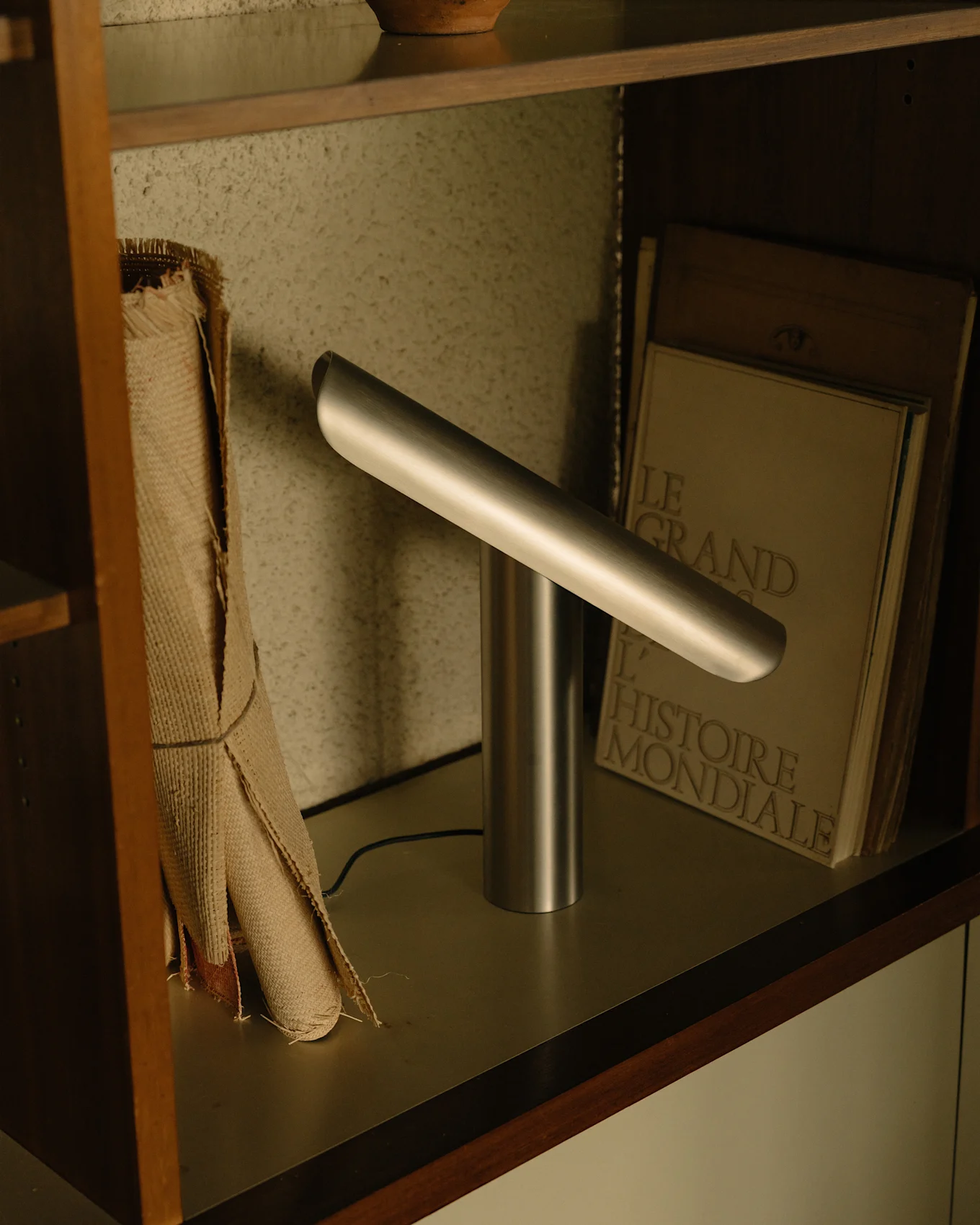 Modern silverfärgad skrivbordslampa med cylindrisk bas och en vinklad horisontell ljusstång på en hylla.