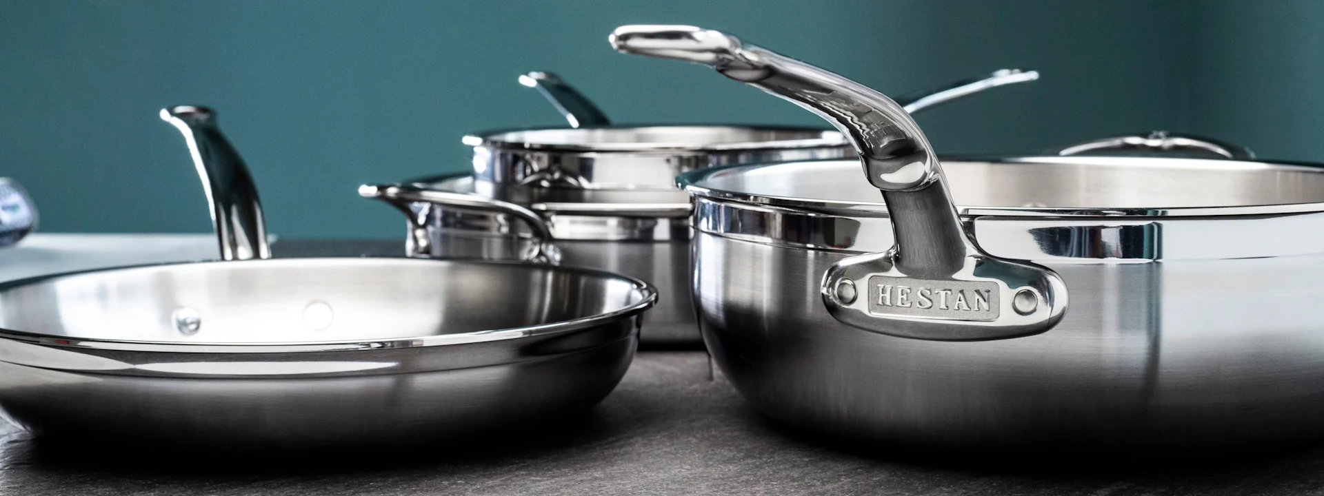 Glänsande grytor och stekpannor i rostfritt stål, inklusive en stor Hestan-märkt panna, på en mörk bänkskiva.