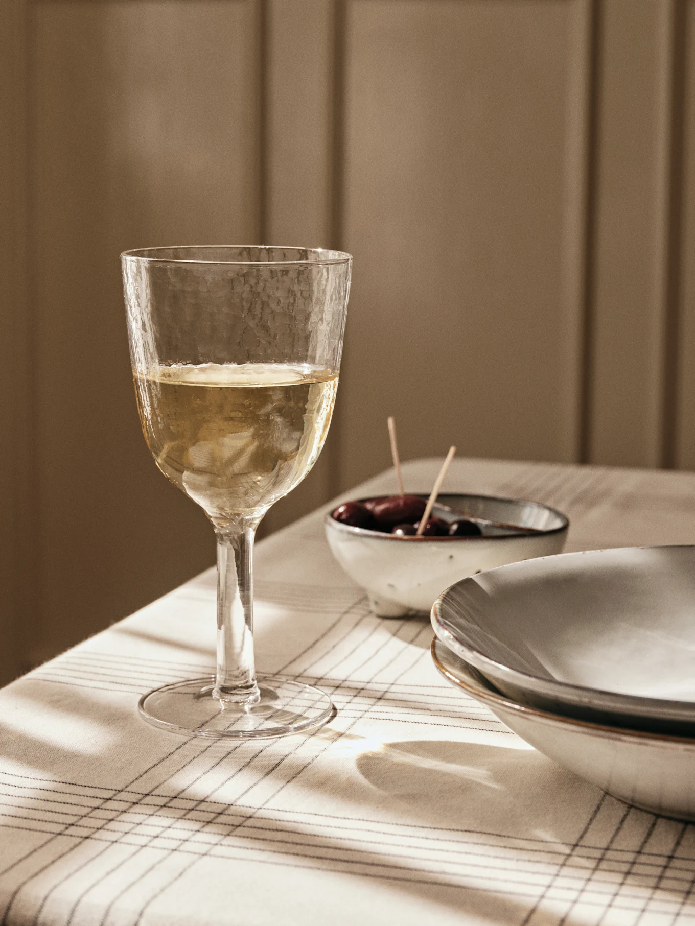 Ett texturerat glas vitt vin på en randig duk, med oliver och tallrikar i bakgrunden, badat i fönsterljus.