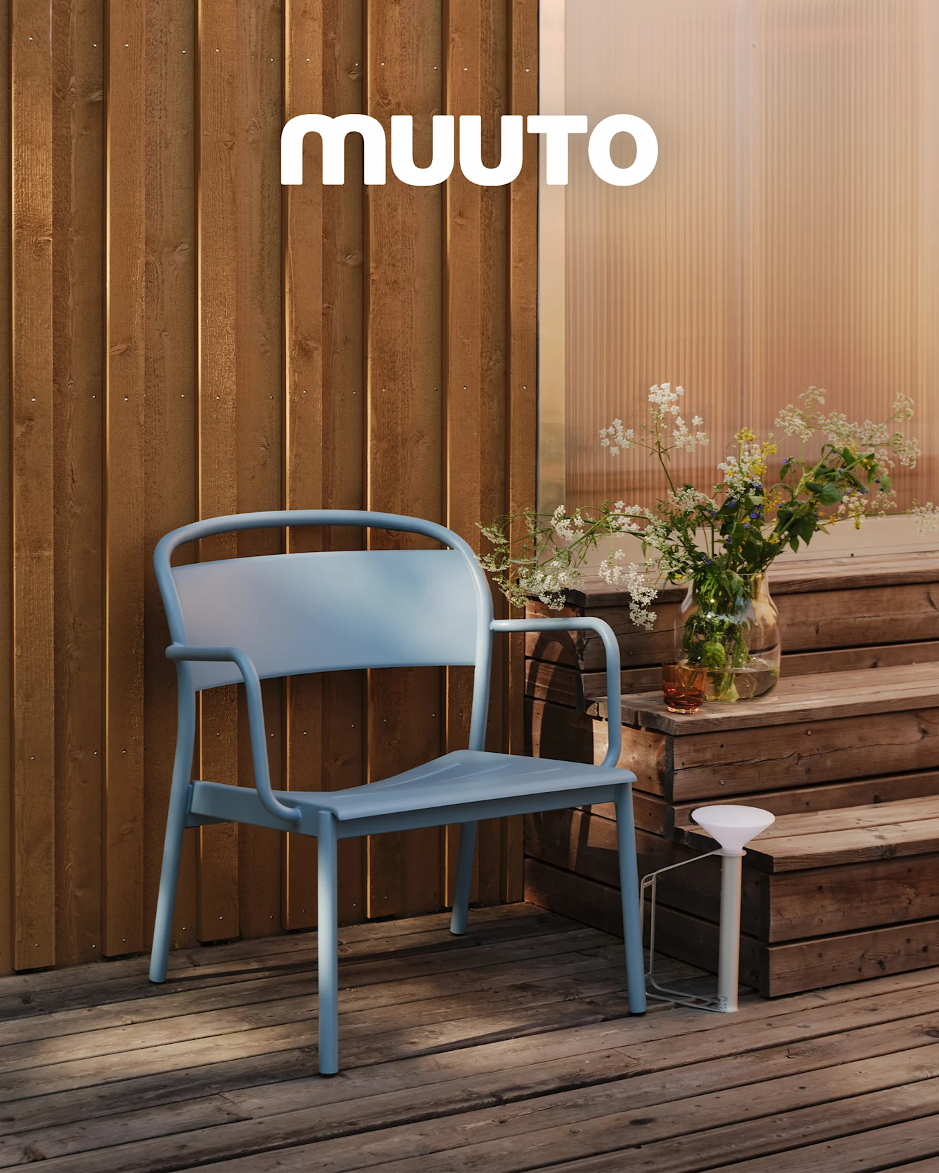 En ljusblå utomhusstol på ett trädäck bredvid trappsteg med en vas vilda blommor och en vit bärbar lampa. MUUTO-logotyp.