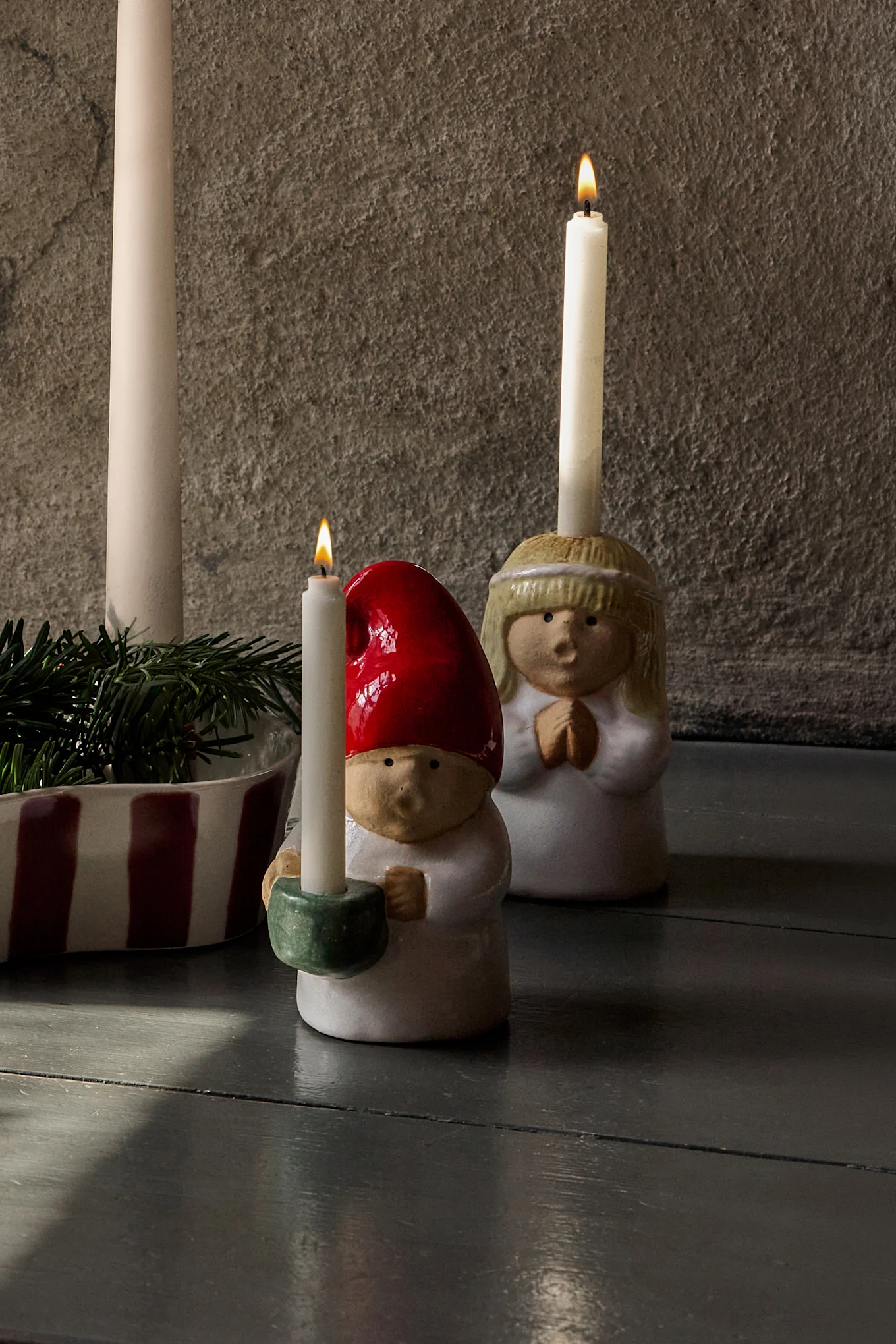Adventsarrangemang: fyra otända ljus i en randig hållare med granris, flankerade av keramiska ängla- och tomtefigurer med tända ljus.