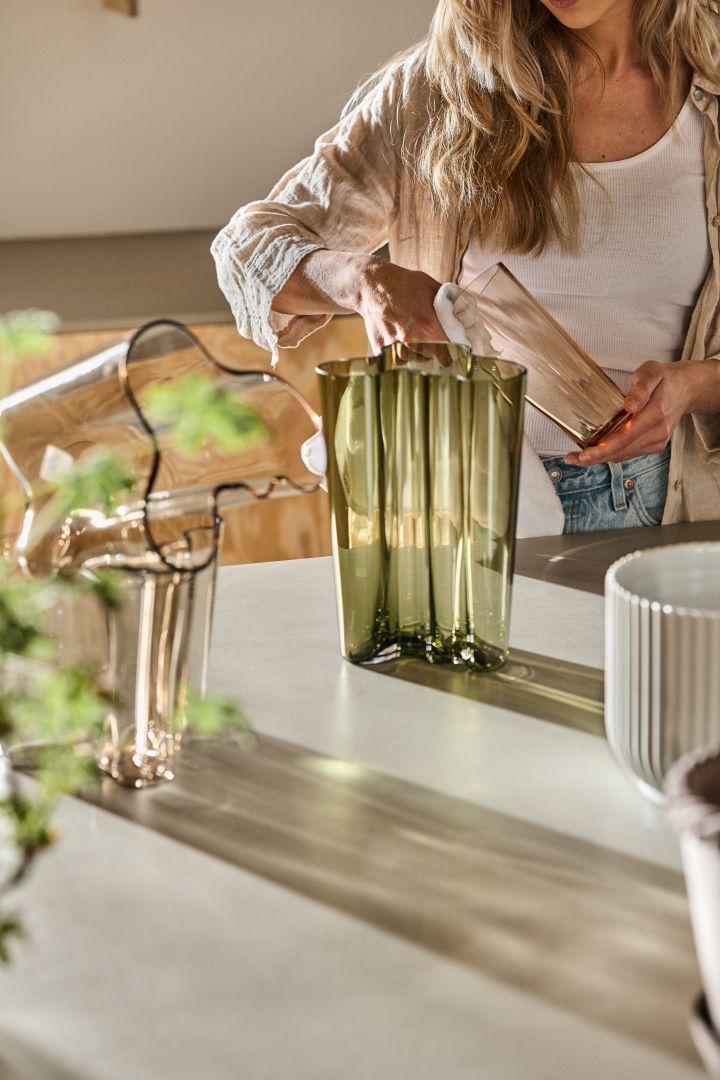 En Alvar Aalto vas i mossgrön står på en köksbänk framför en kvinna som putsar en annan Alvar Aalto vas. 