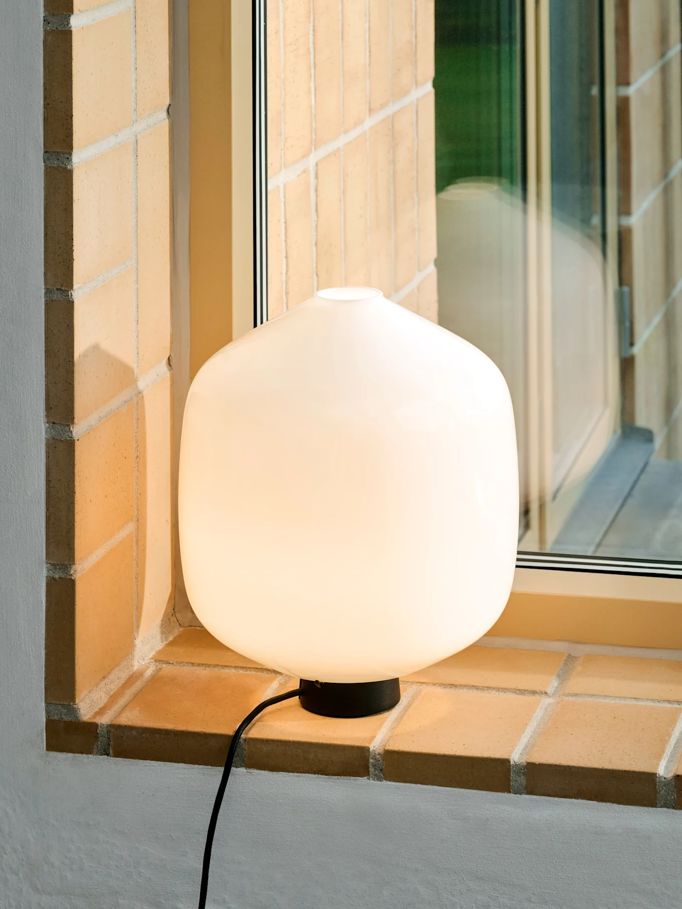 En glödande, rundad vit bordslampa med svart bas och sladd på en tegelfönsterbräda.