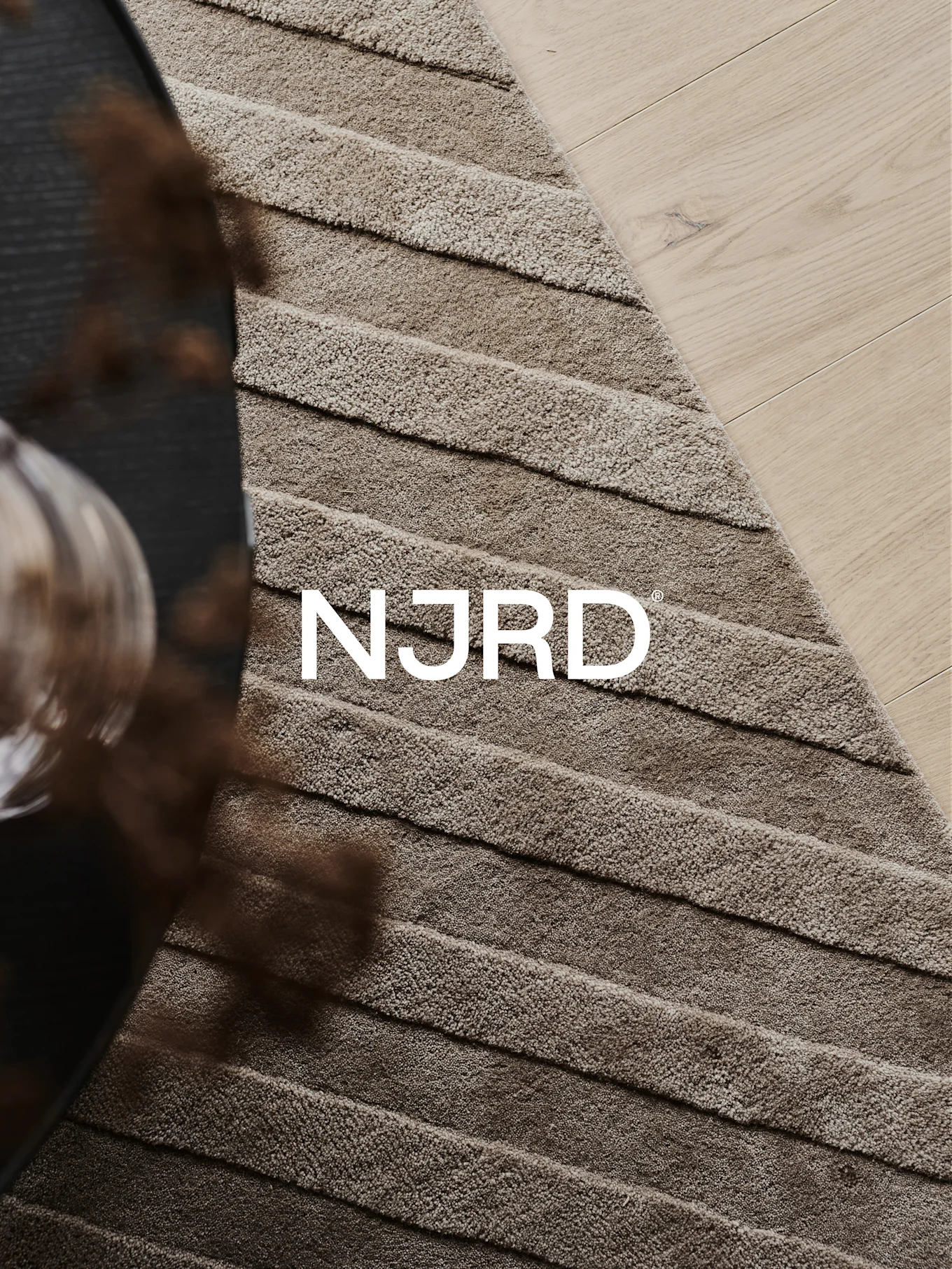 Närbild av en ljusbrun ryamatta med horisontella räfflor, delvis på ett ljust trägolv, med den vita 'NJRD'-logotypen.