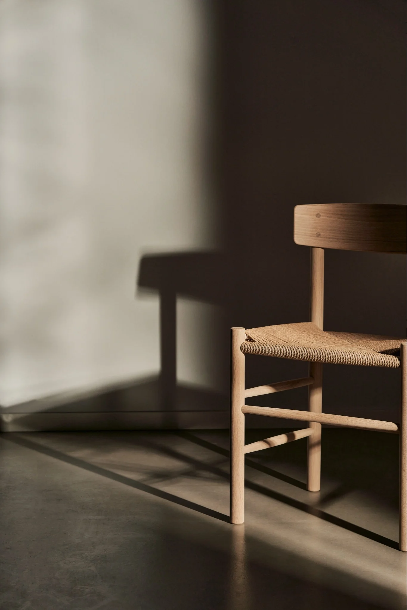 En stol i bok med rena linjer och vackert hantverk står ensam i ett tomt rum, där den kastar en mjuk skugga mot väggen bakom.