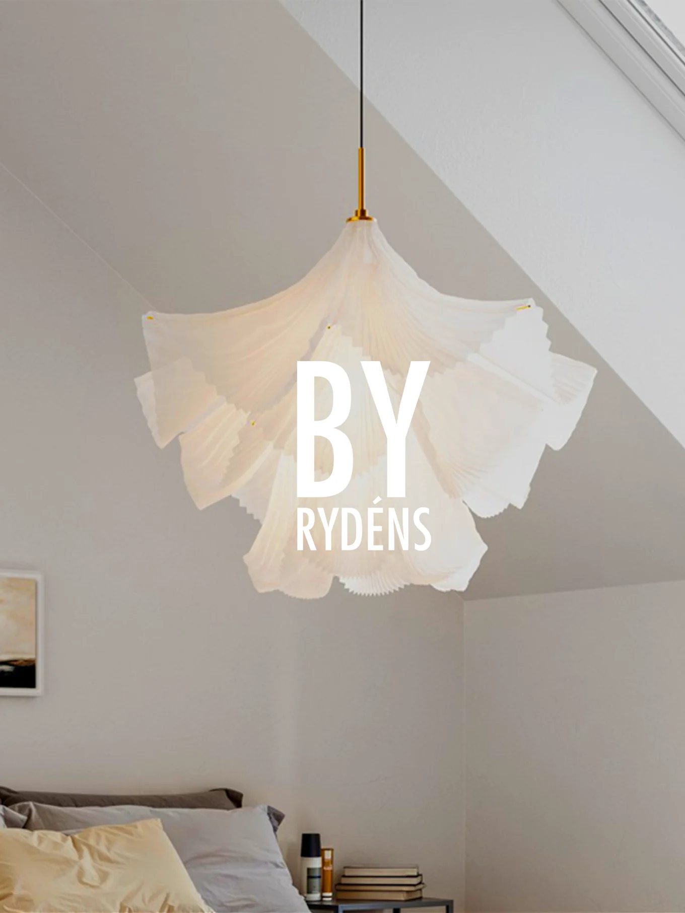 En vit, veckad skulptural taklampa hänger ovanför en säng med kuddar och ett sängbord. 'BY RYDÉNS' är överlagrat på lampan.