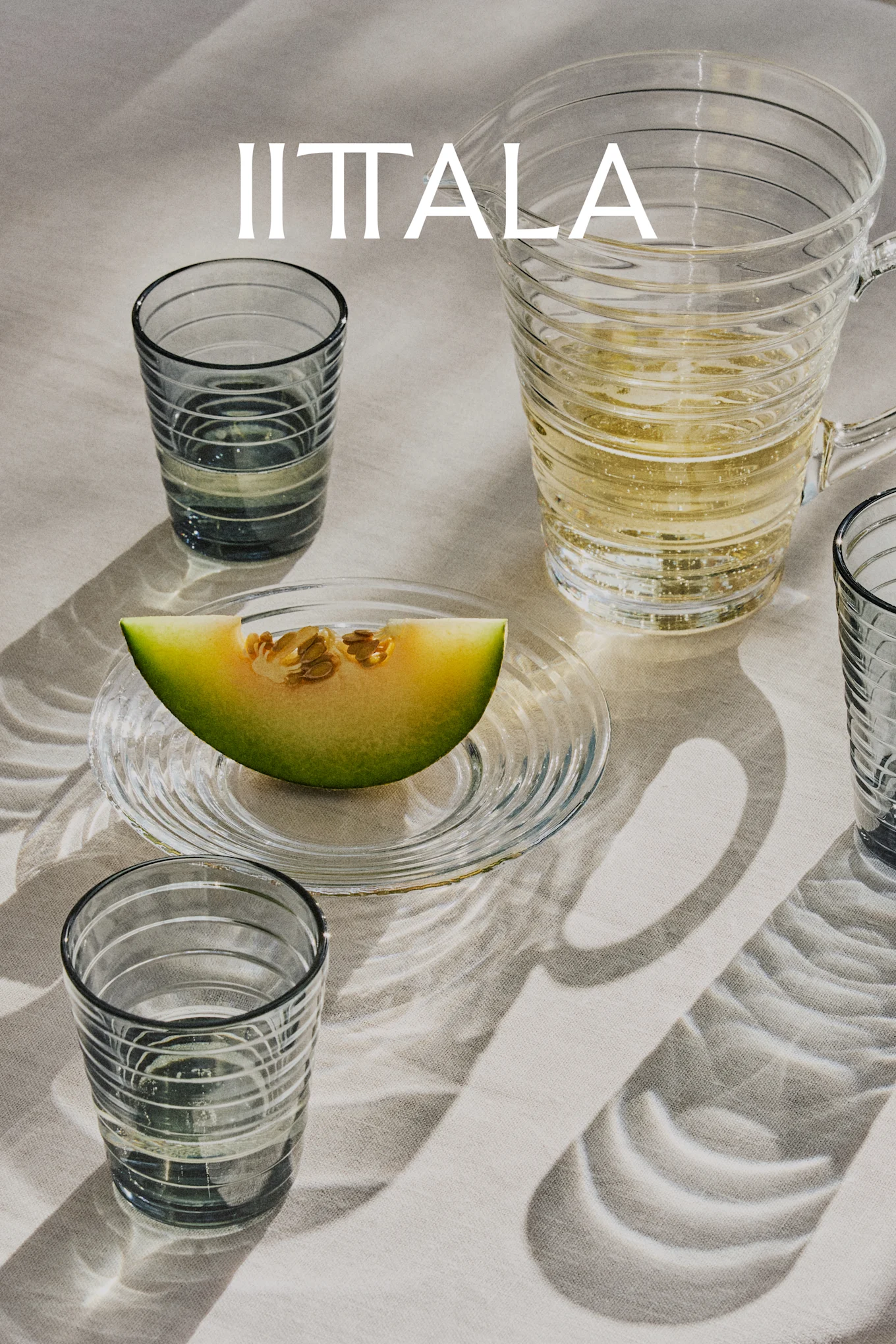 IITTALA-logga över glas, en kanna med bubbeldryck och en melonskiva på fat, allt på ljust tyg.