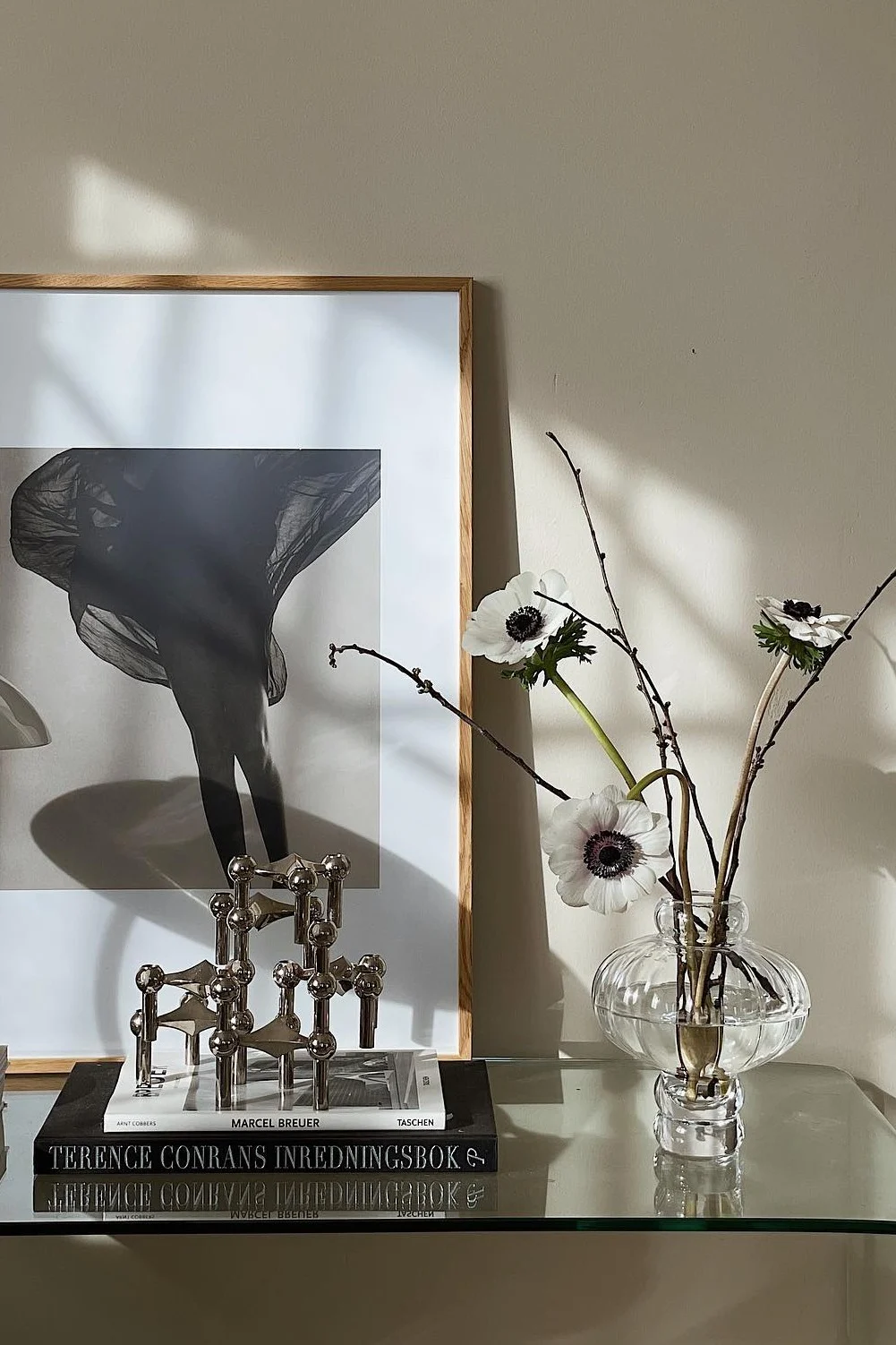 Inramad svartvit bild av ben med flödande tyg, modulär ljusstake, böcker och vita anemoner i vas.