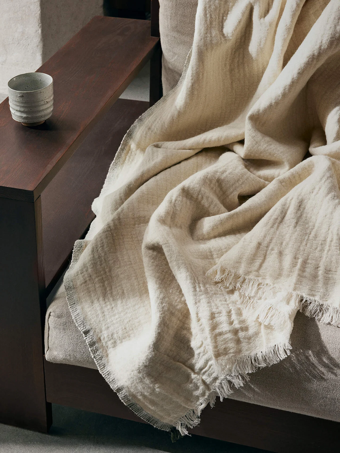 En ljusbeige texturerad filt med fransade kanter draperad över en soffa, med en keramisk kopp på ett mörkt träarmstöd.