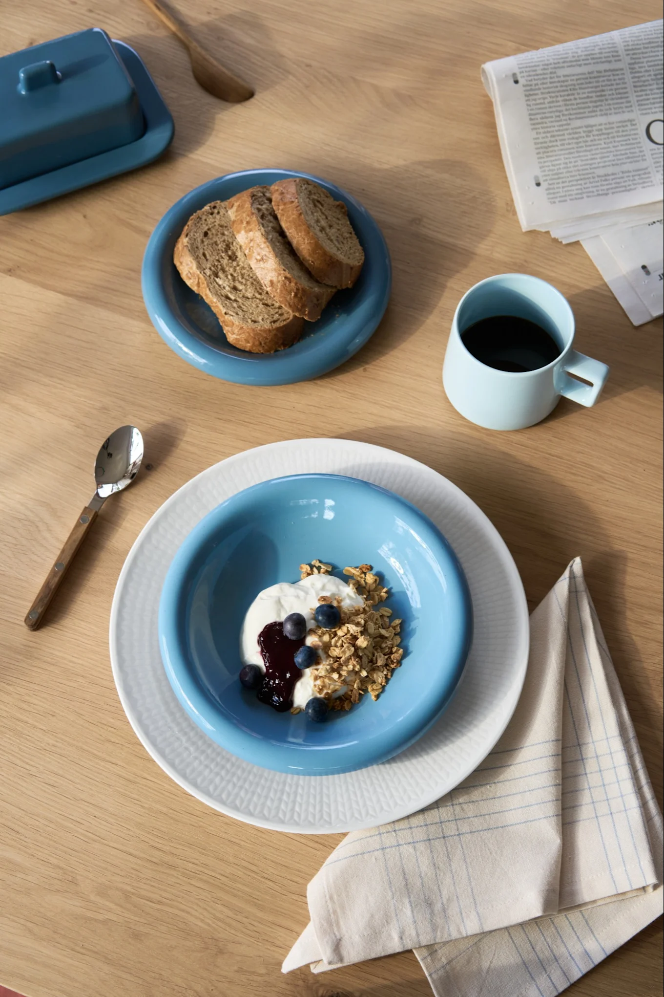 Frukost med yoghurt, granola, blåbär och bröd på blå och vita tallrikar, kaffe i blå mugg, smörask på träbord.