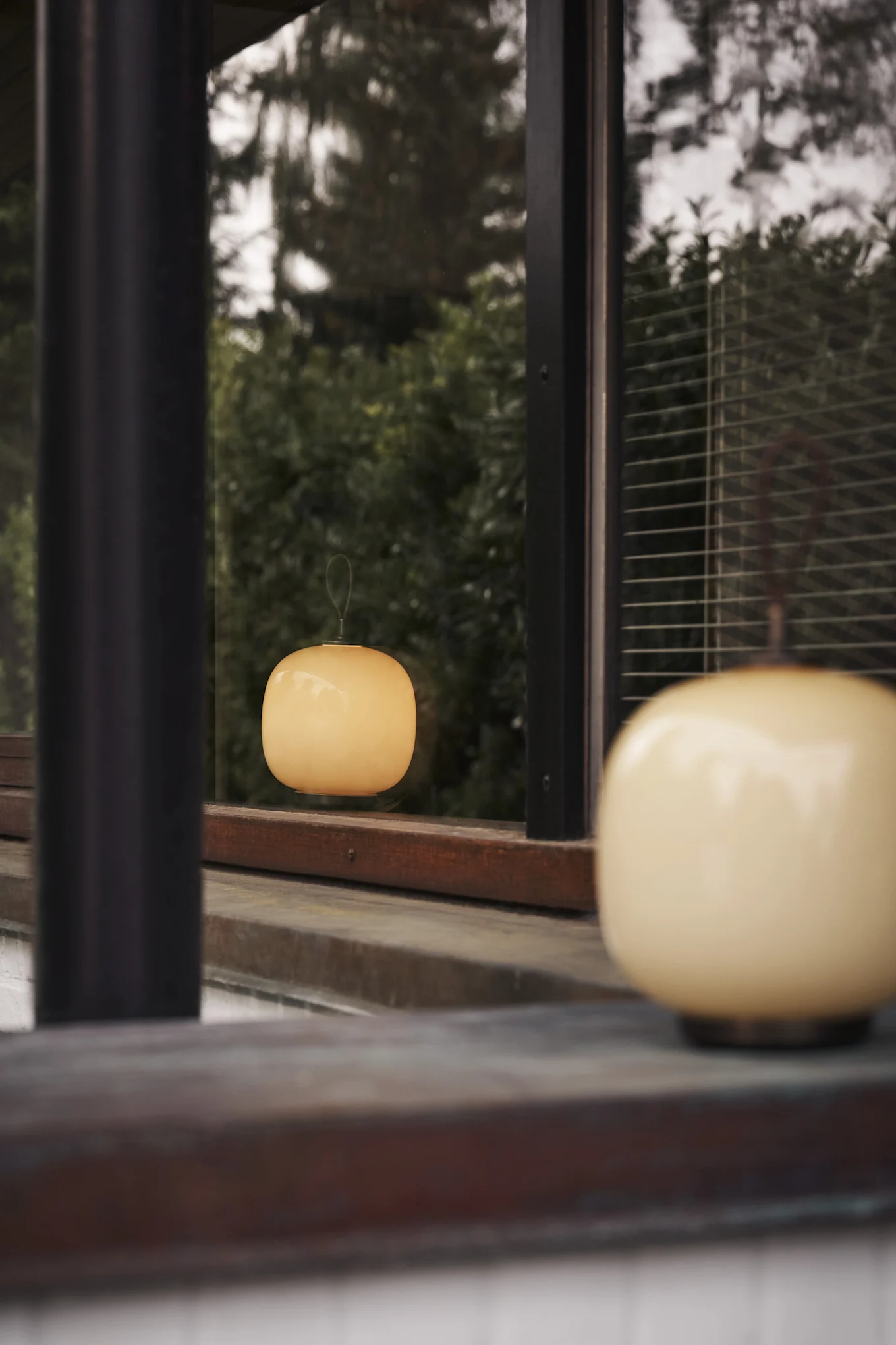 Två ljusa, runda lampor, en suddig i förgrunden och en reflekterad i ett fönster med grönt lövverk. Inspiration till din balkong 2026.