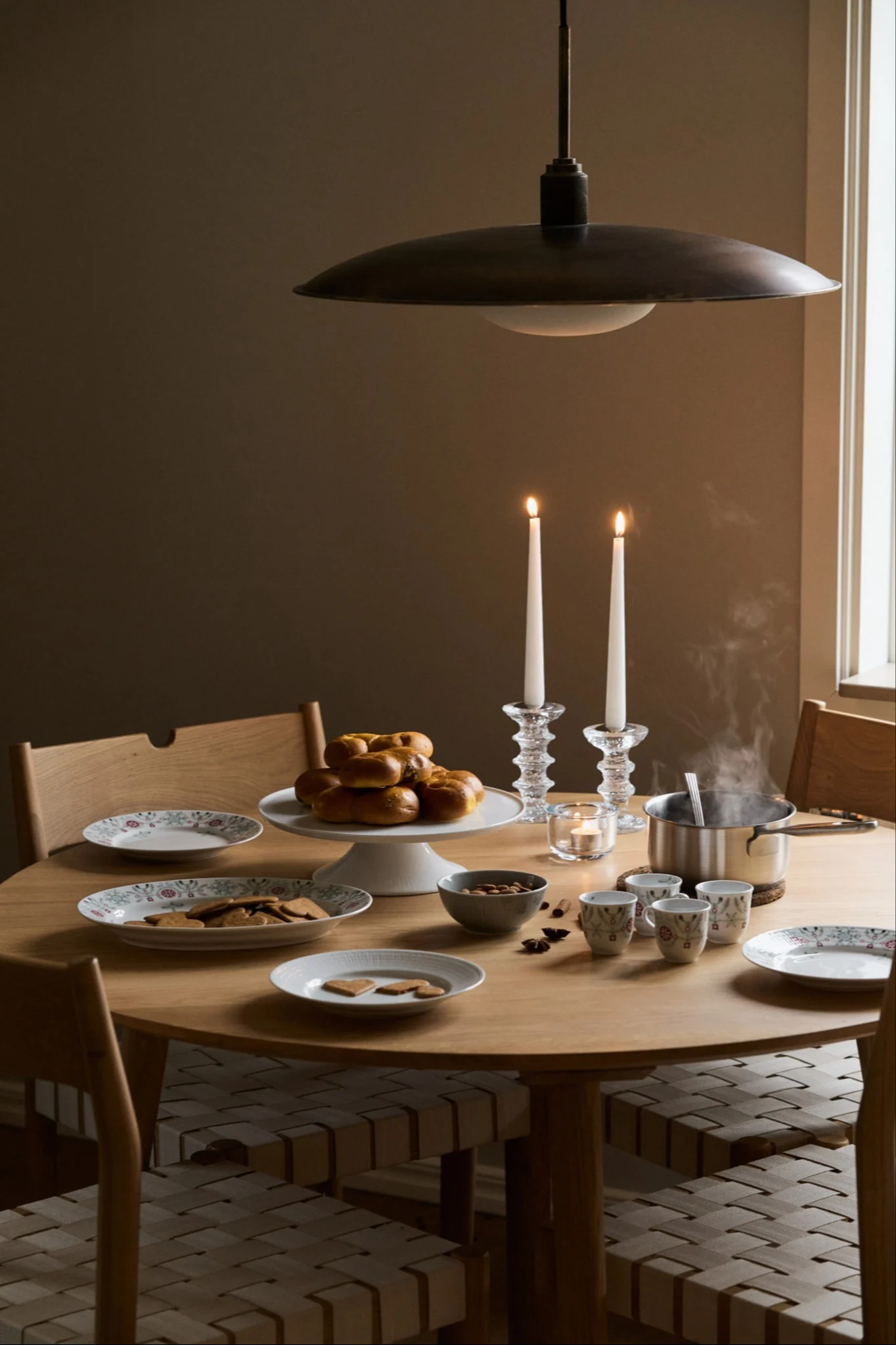Ett träbord dukat med servisen Swedish Grace Winter från Rörstrand inför första advent. På bordet står ett kakfat med lussekatter, en tallrik med pepparkakor, en kastrull med varm dryck, små koppar och två tända ljus i glasljusstakar.