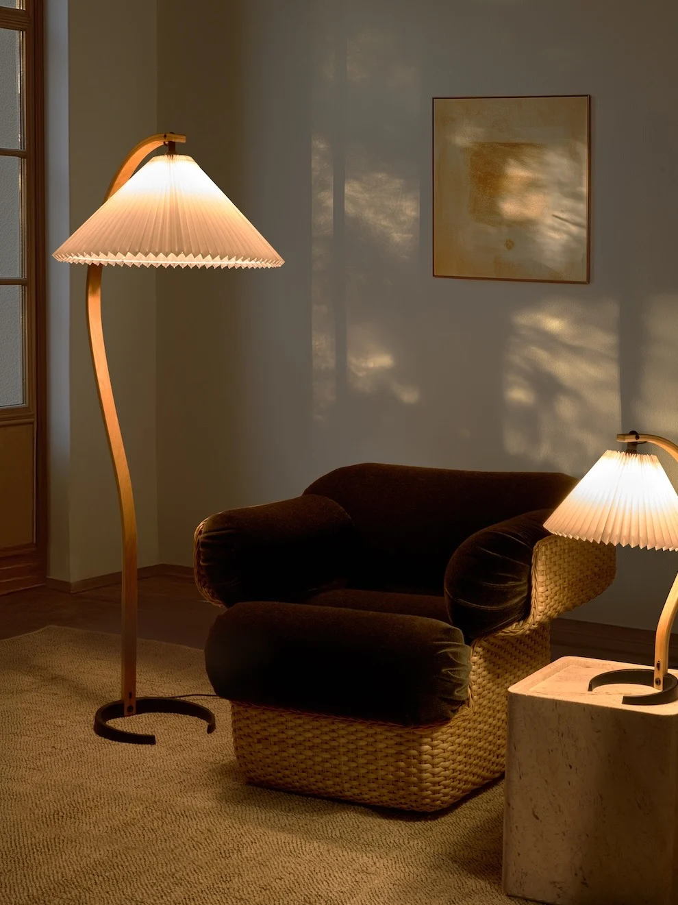 Mysigt rum med en böjd golvlampa, mörk fåtölj med flätad bas och en mindre bordslampa. Varmt ljus skapar skuggor.