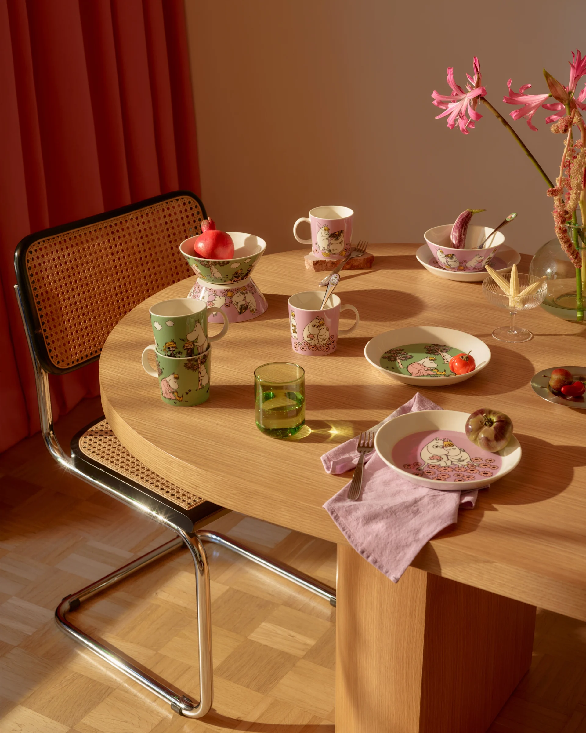 Matbord med rosa och gröna Mumin-serviser, färsk frukt, ett grönt glas och rosa blommor.