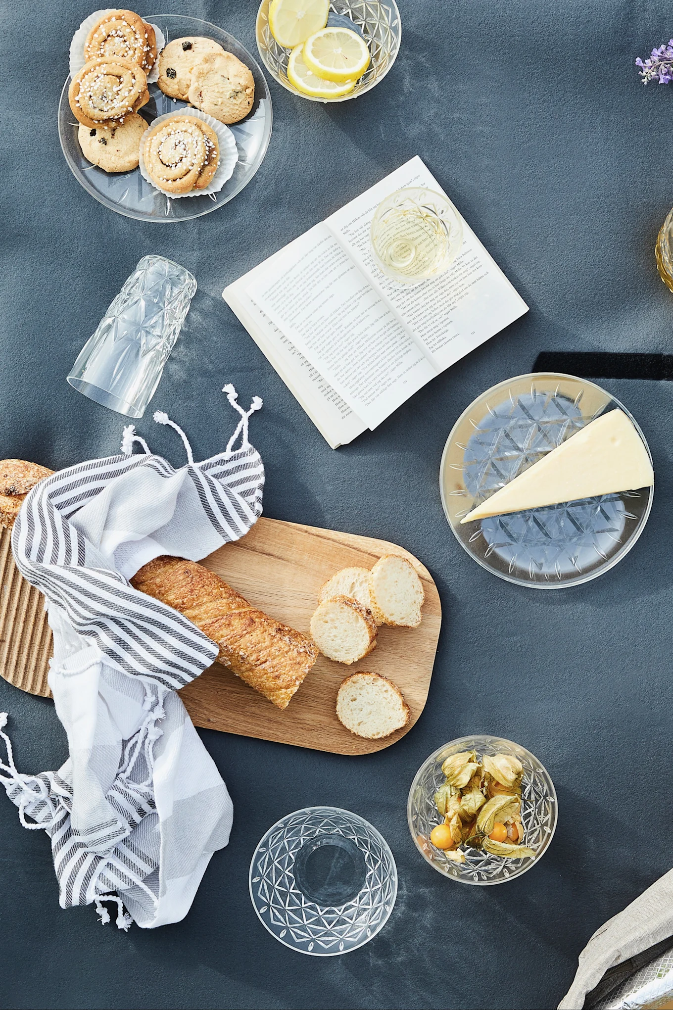 Översiktsbild av en picknick med kakor, citroner, vin, ost, baguette, physalis och en öppen bok på en grå filt.