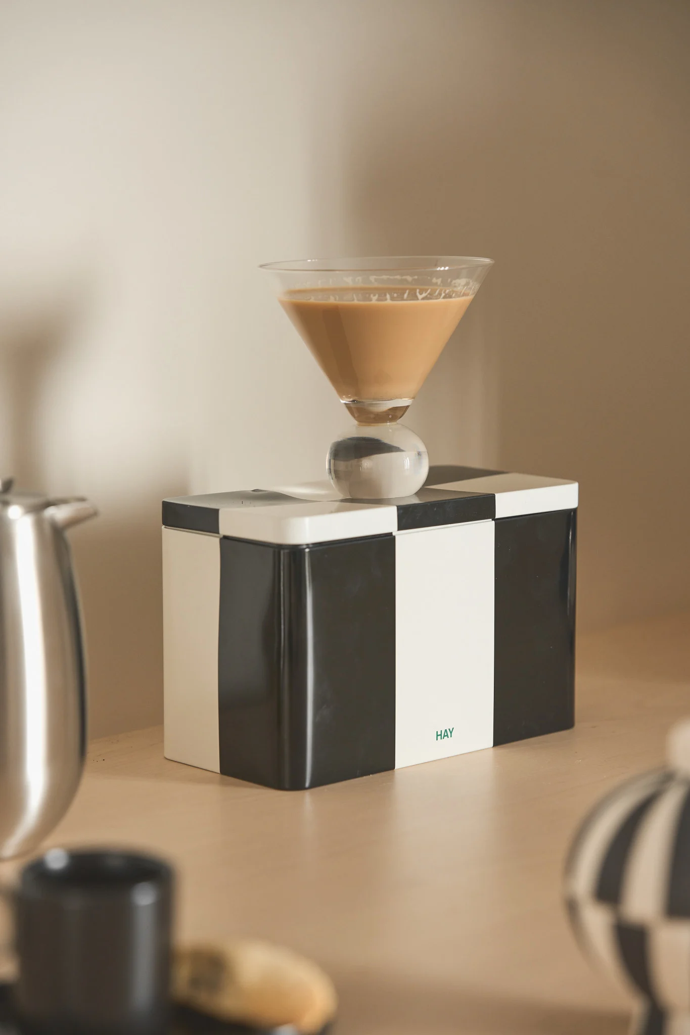 Martiniglas med ljusbrun dryck och sfärisk fot på en svart- och vitrutig låda med 'HAY'-logga.
