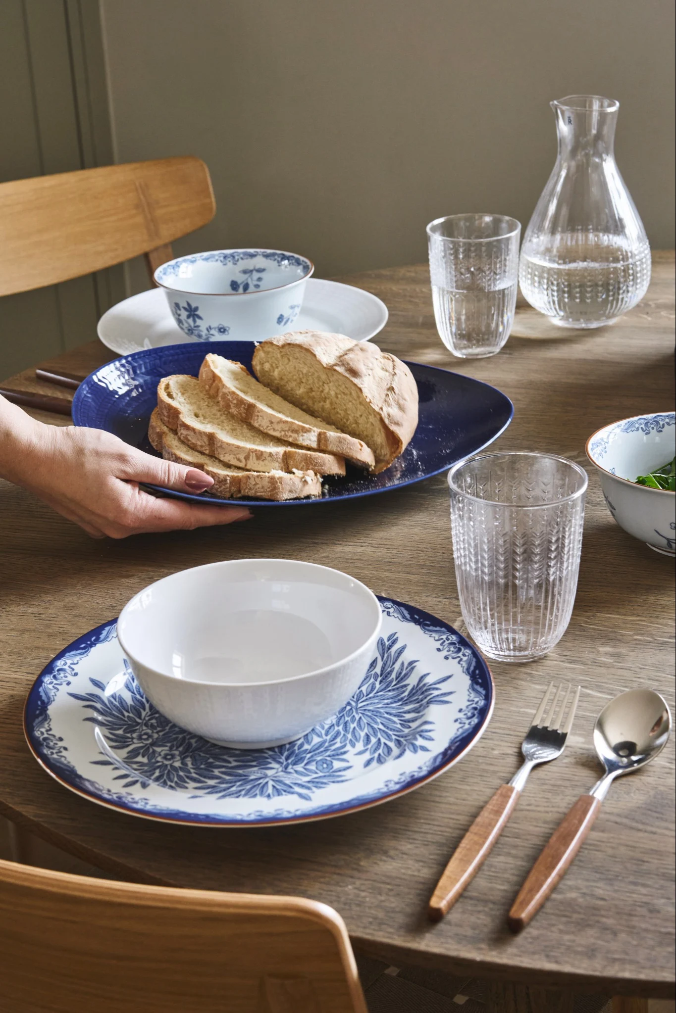 Händer placerar ett blått fat med skivat bröd på ett bord dukat med blåvita tallrikar, glas och bestick från Rörstrand.