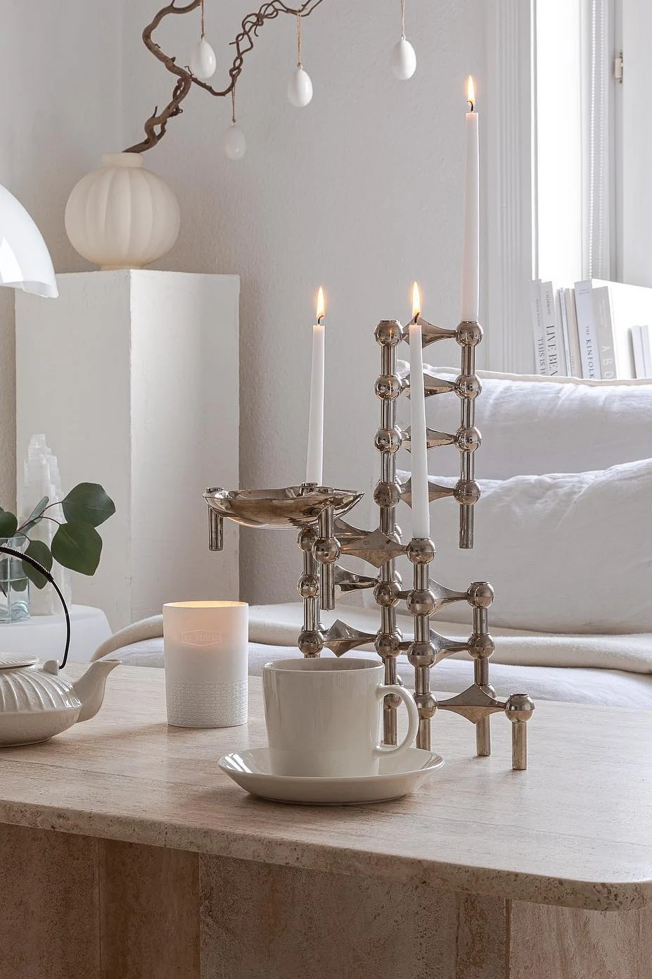 Modern silverfärgad ljusstake med tre tända vita ljus, tekopp och tekanna på stenbord med påskdekoration.