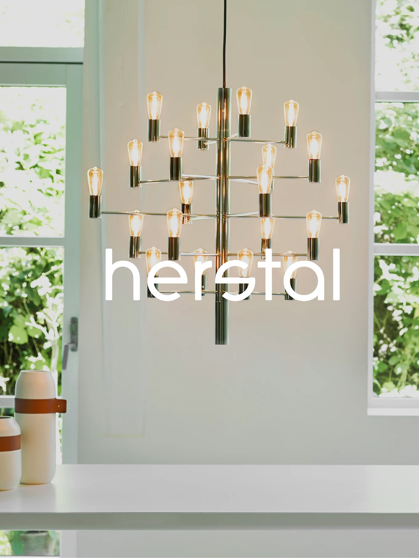 Herstal ljuskrona med flera synliga glödlampor i ett ljust rum med stora fönster.