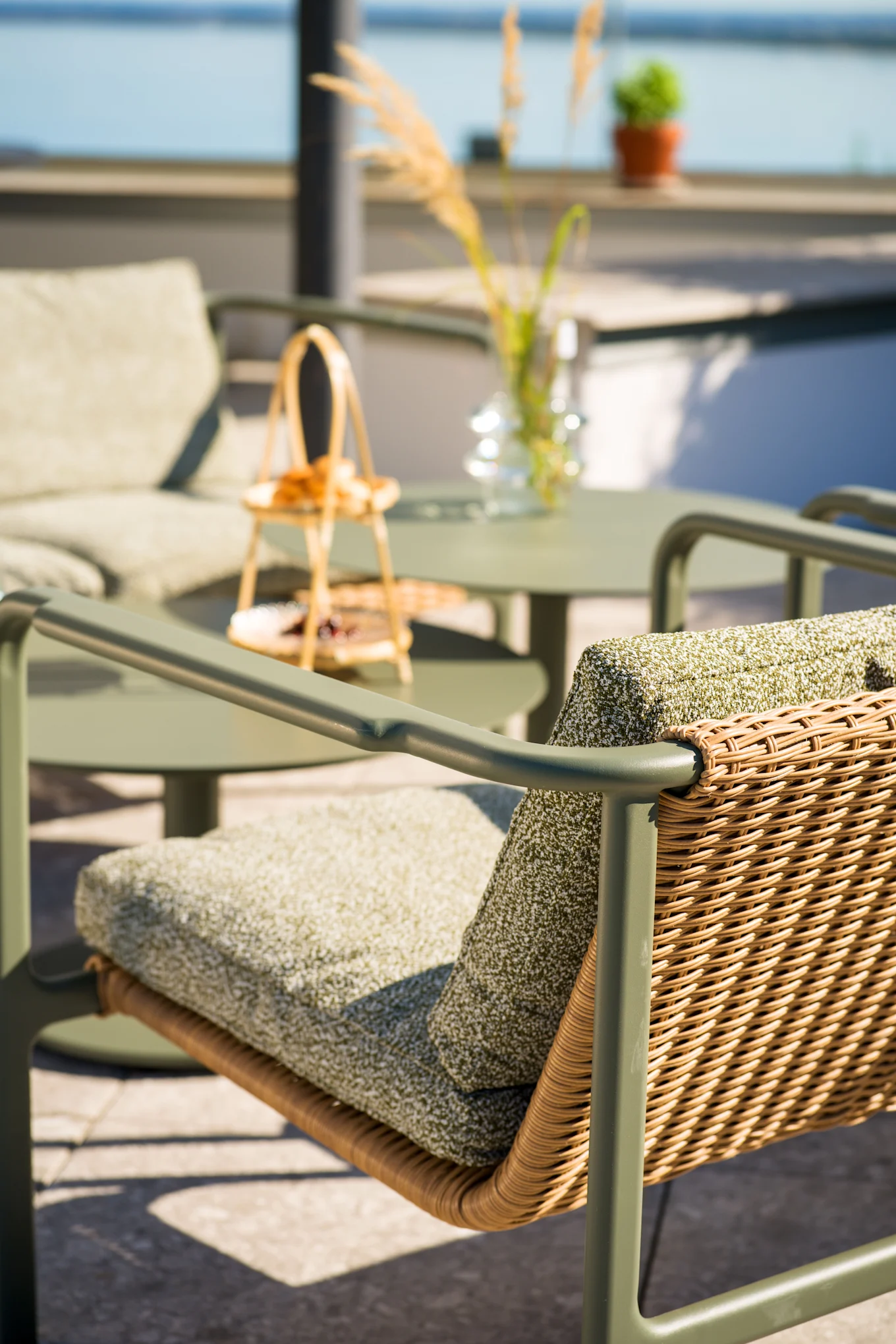 Utomhusterrassstol med olivgrön ram, flätad rottingrygg och gröna dynor. Matchande bord och stol i suddig bakgrund nära vatten.