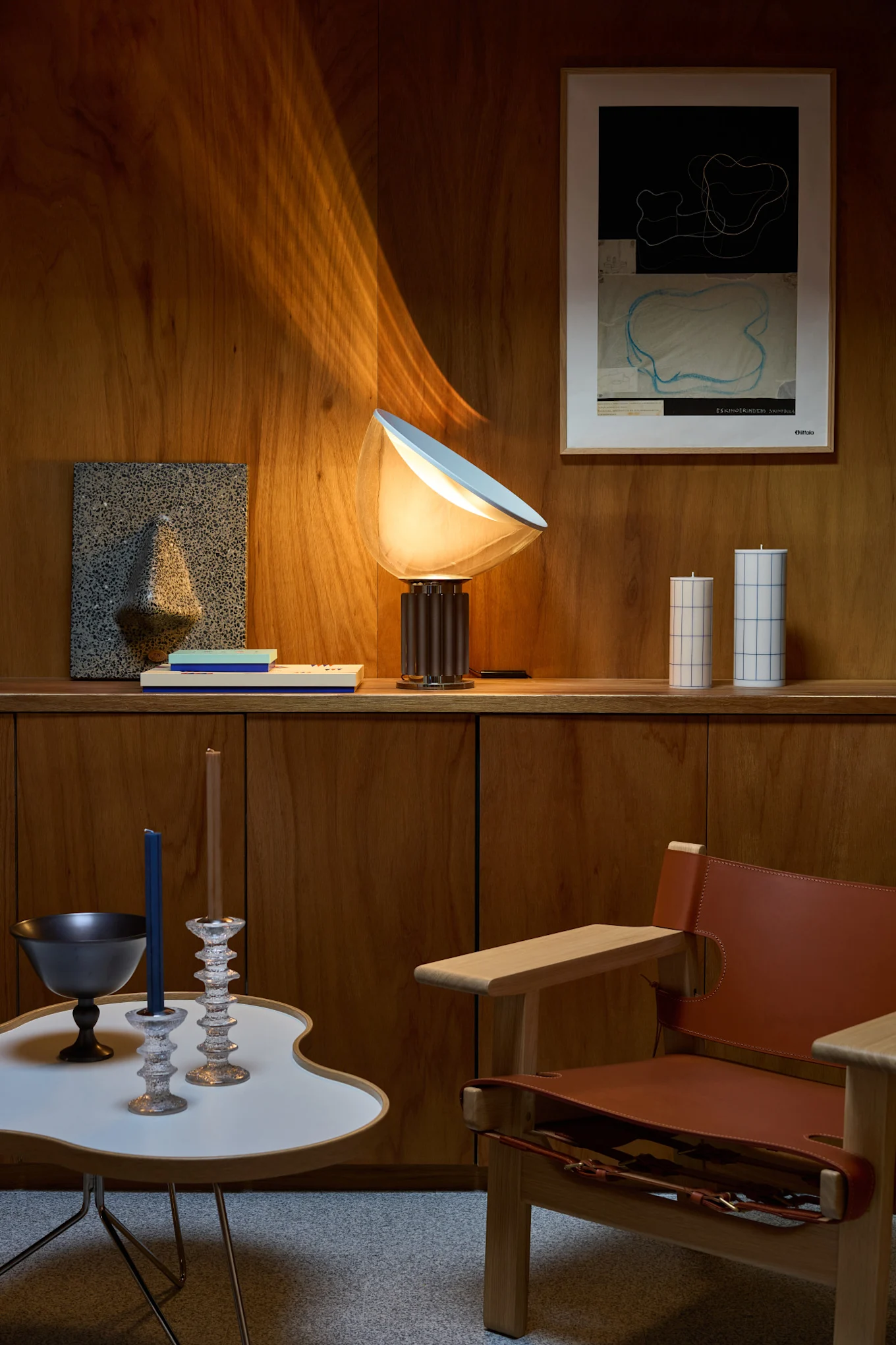 Ett vardagsrum med träväggar, en glödande bordslampa, abstrakt väggkonst, dekorativa ljus, ett modernt sidobord och en läderfåtölj.