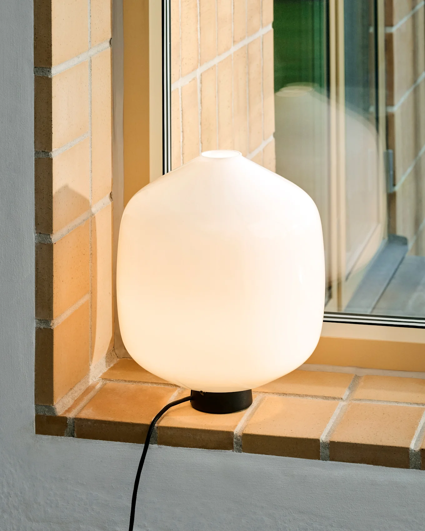En glödande vit rundad bordslampa med svart bas och sladd står på en ljus tegelstensfönsterbräda.