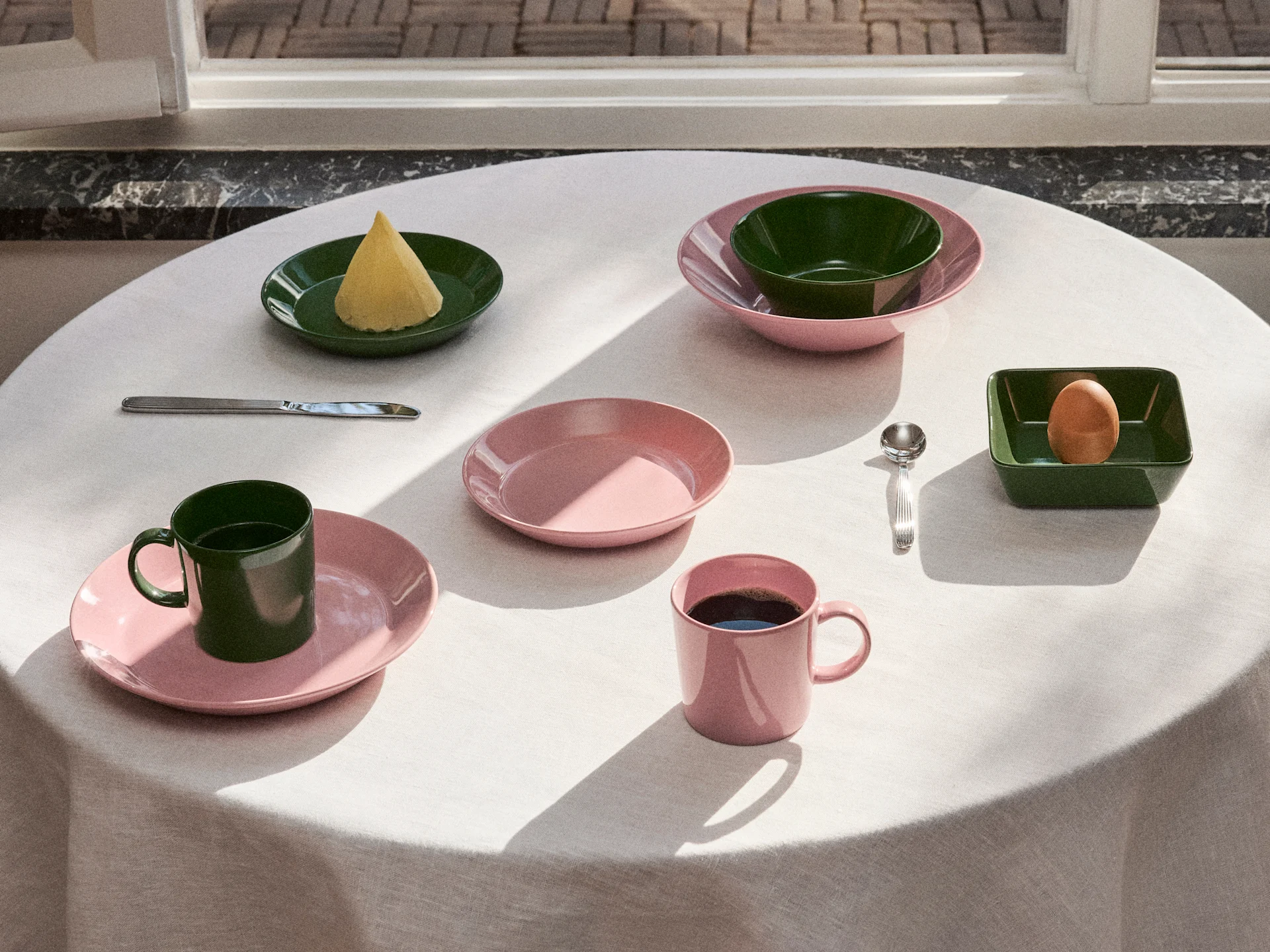 Bord dukat med rosa och mörkgrönt porslin, tallrikar, skålar och muggar, med smör, ett ägg och kaffe i naturligt ljus.