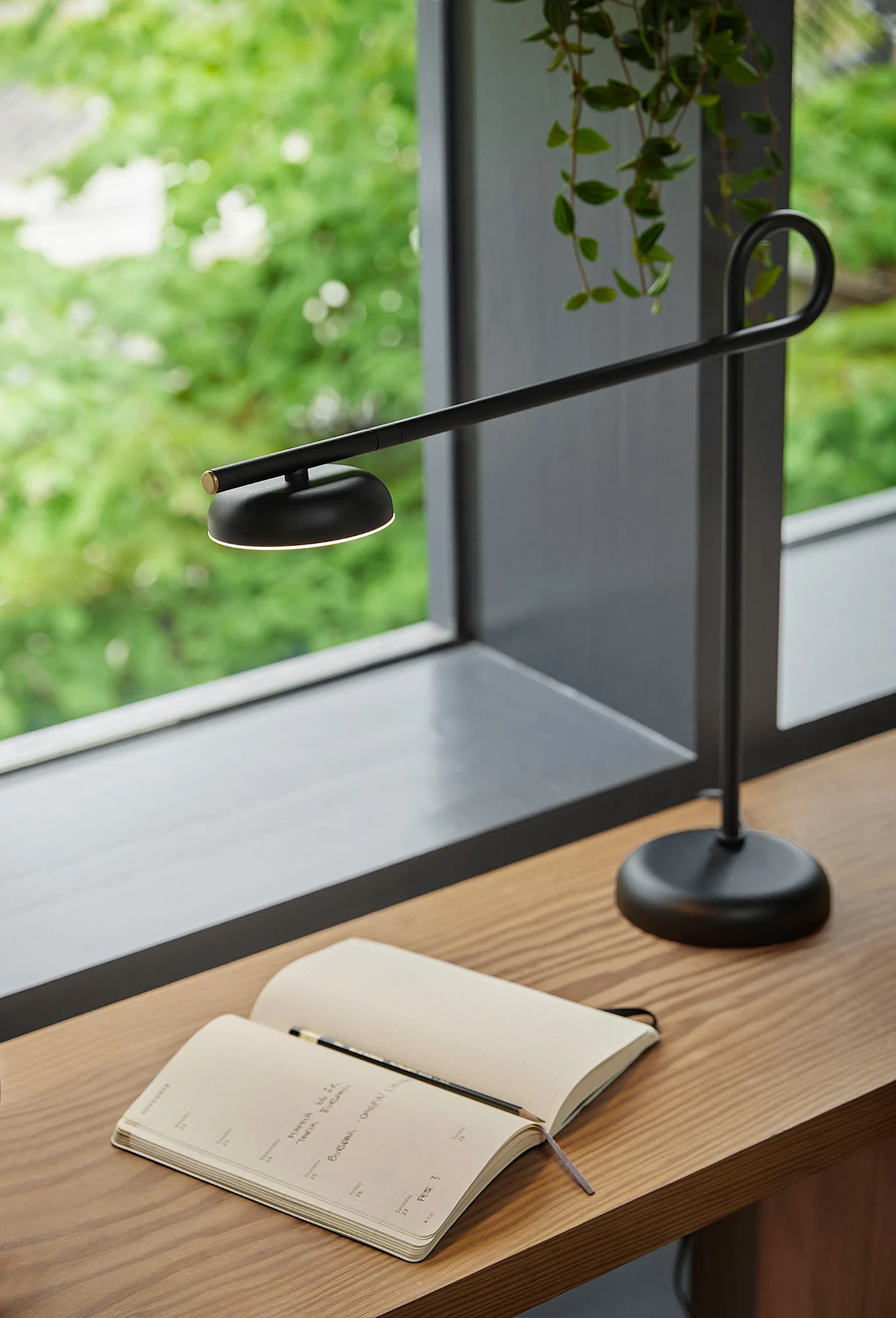 En modern svart skrivbordslampa lyser upp en öppen anteckningsbok och penna på ett träskrivbord bredvid ett fönster med gröna löv.