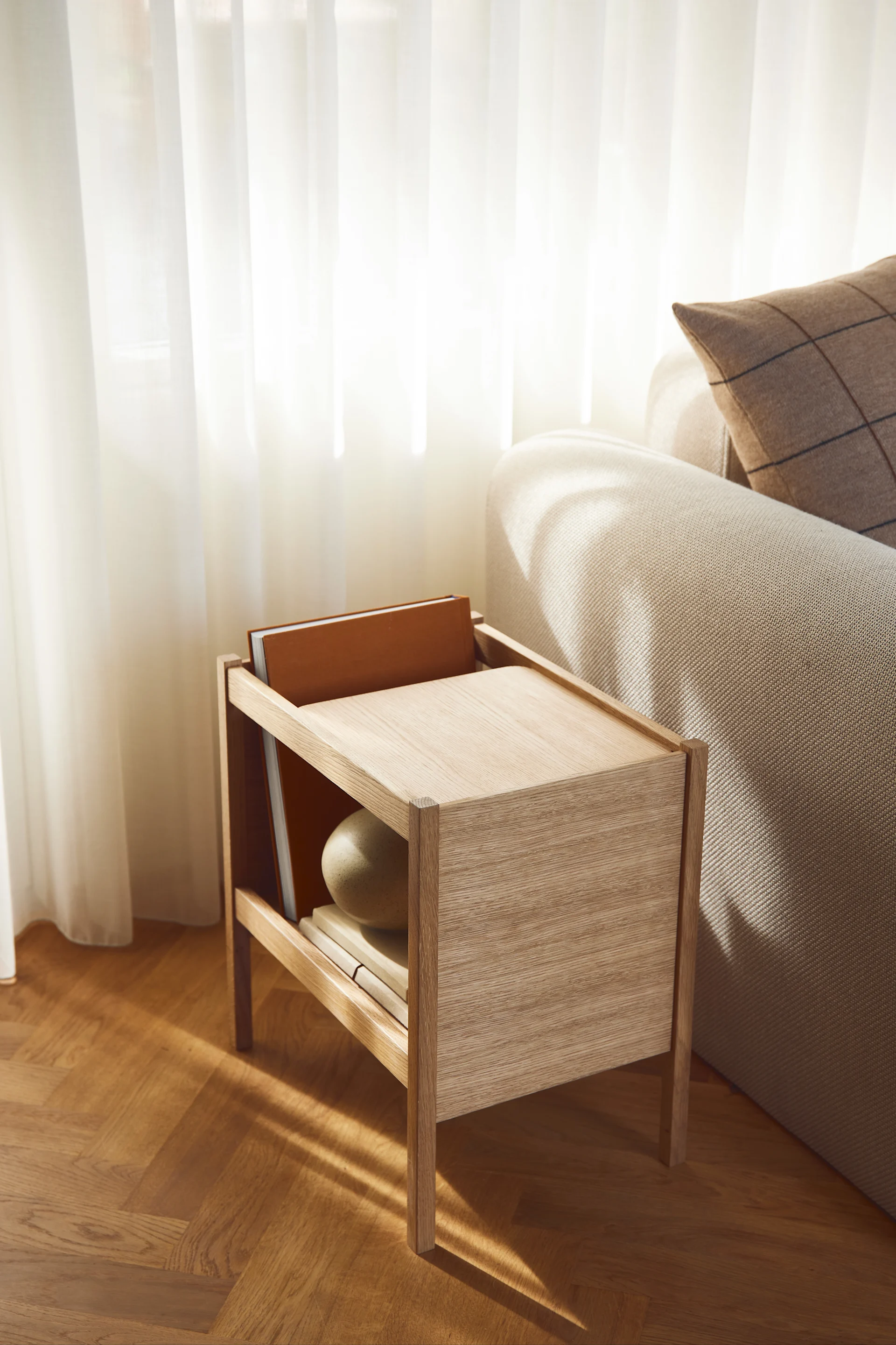 Ett ljust träbord med böcker och en dekorativ sfär bredvid en beige soffa, badat i solljus från skira gardiner.