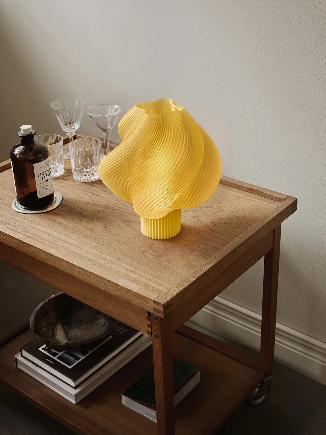 Gul plisserad lampa på rullande träbord med ginflaska, glas och böcker på nedre hyllan.