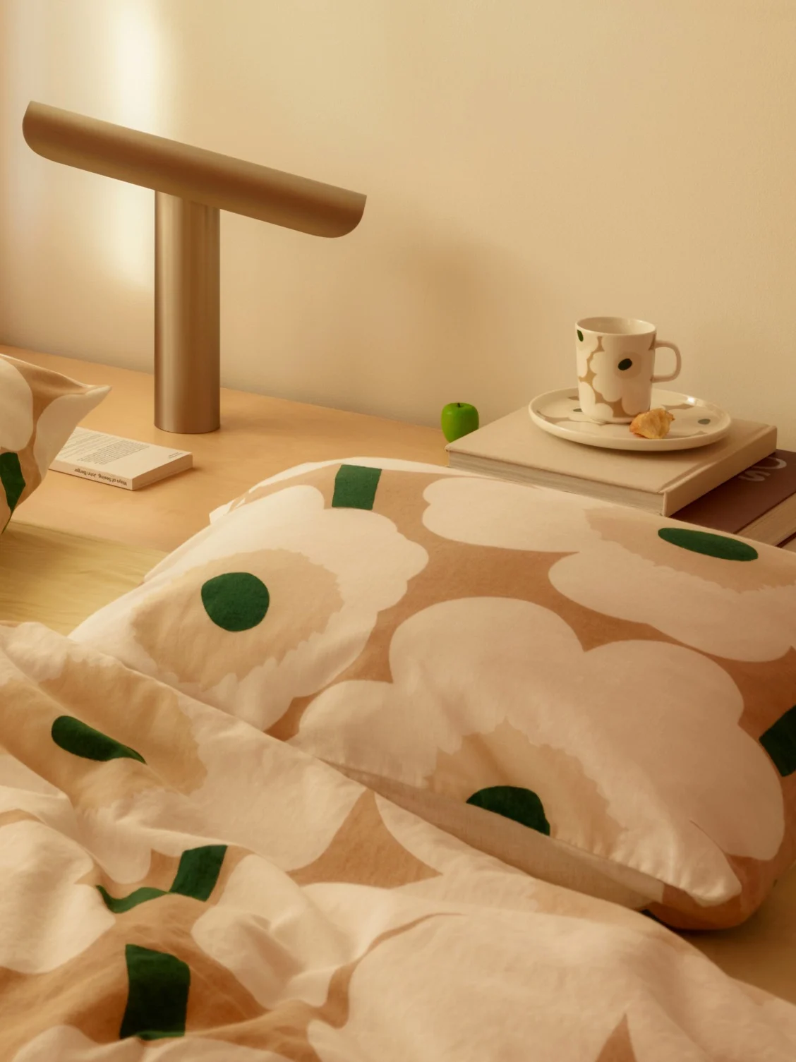Sängbild med vit och beige blommig sängkläder, en modern lampa, en kopp och en bok på ett nattduksbord i trä.