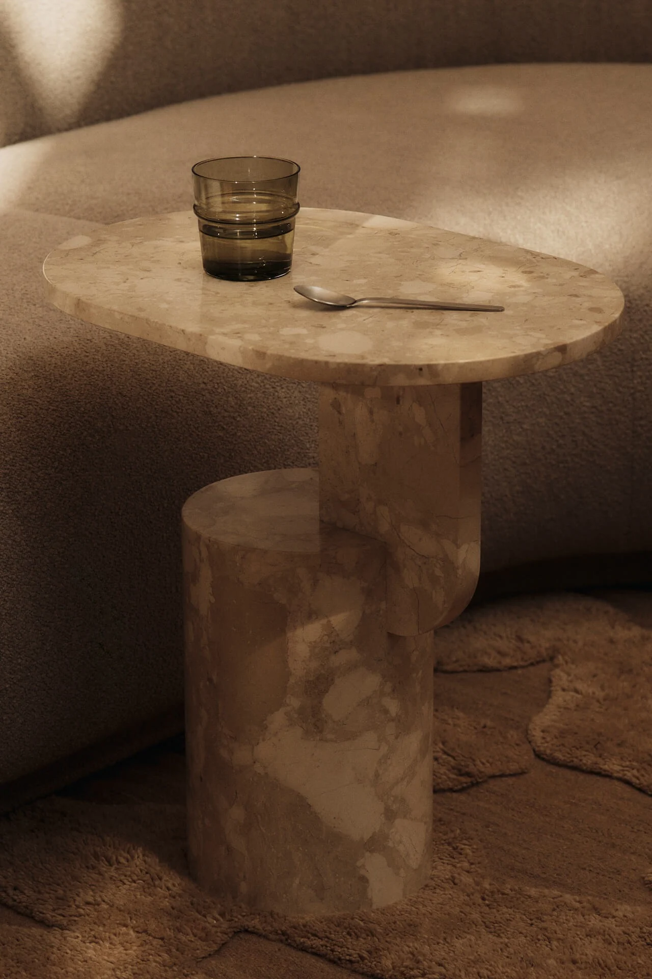 Beigt marmorbord med ett mörkgrönt glas vatten och en silversked, bredvid en texturerad soffa och matta.